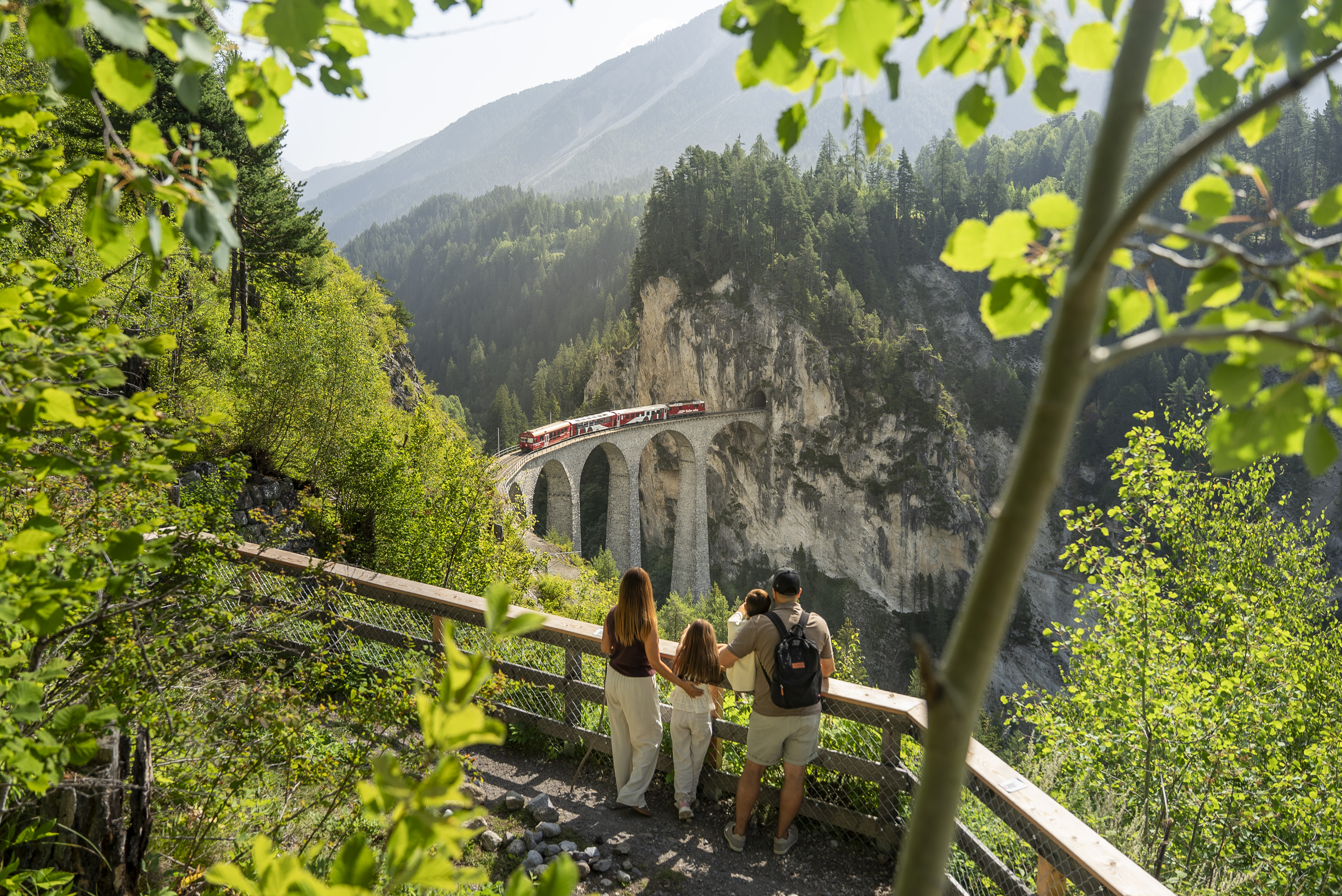 Eine Familie auf der Hennings Aussichtsplattform. Im Hintergrund fährt ein roter Zug über das Landwasser-Viadukt.