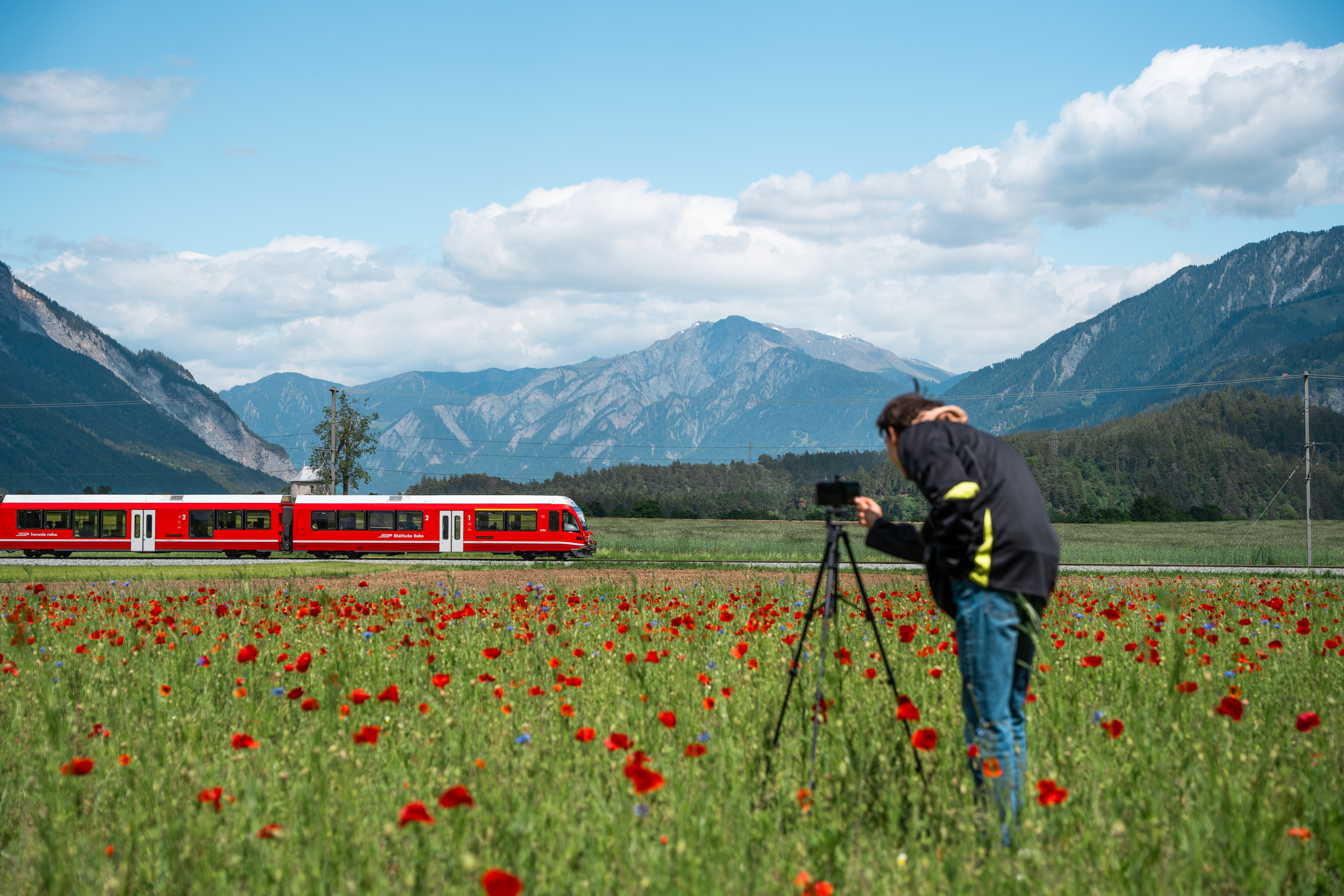Ein junger Mann steht hinter seinem Stativ mit Smartphone und filmt einen vorbeifahrenden, roten Zug der RhB.