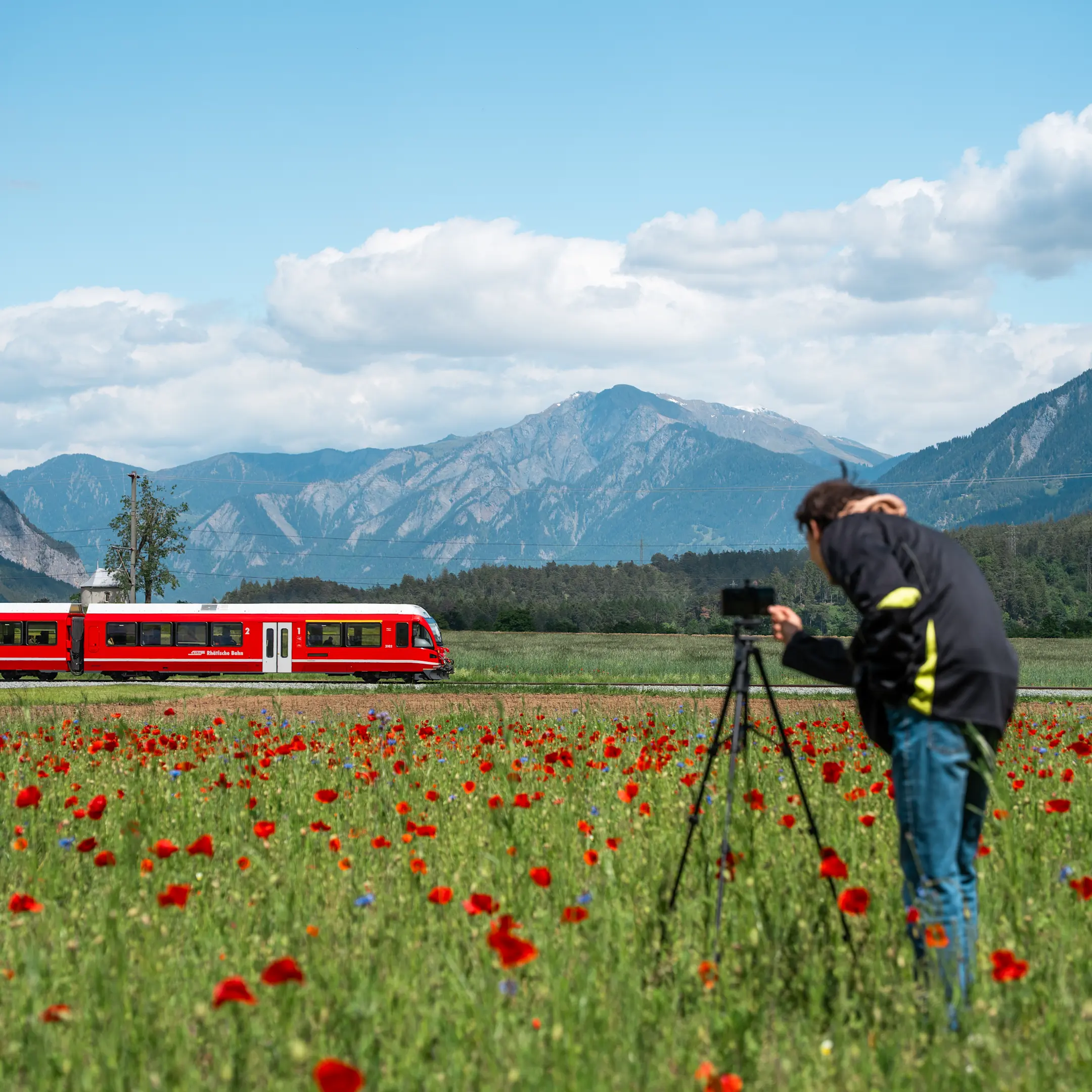 Ein junger Mann steht hinter seinem Stativ mit Smartphone und filmt einen vorbeifahrenden, roten Zug der RhB.