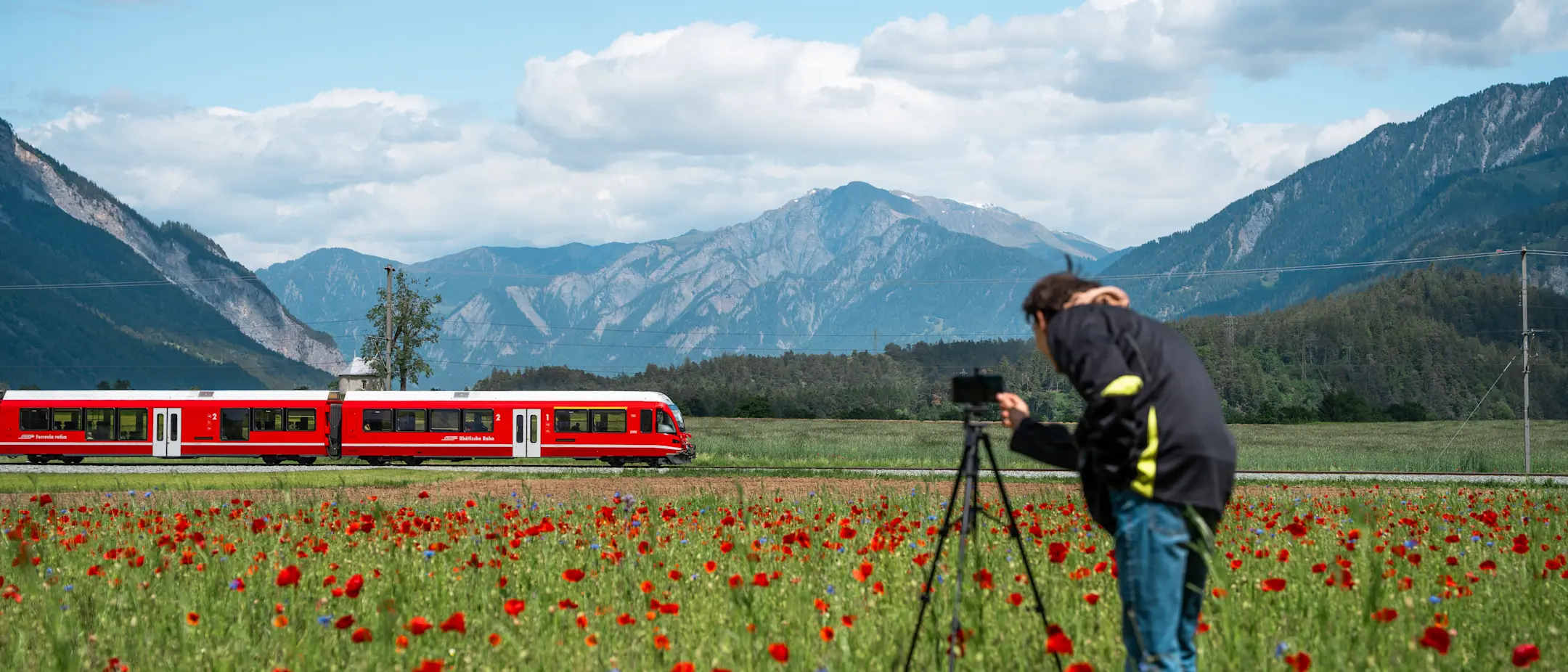Ein junger Mann steht hinter seinem Stativ mit Smartphone und filmt einen vorbeifahrenden, roten Zug der RhB.
