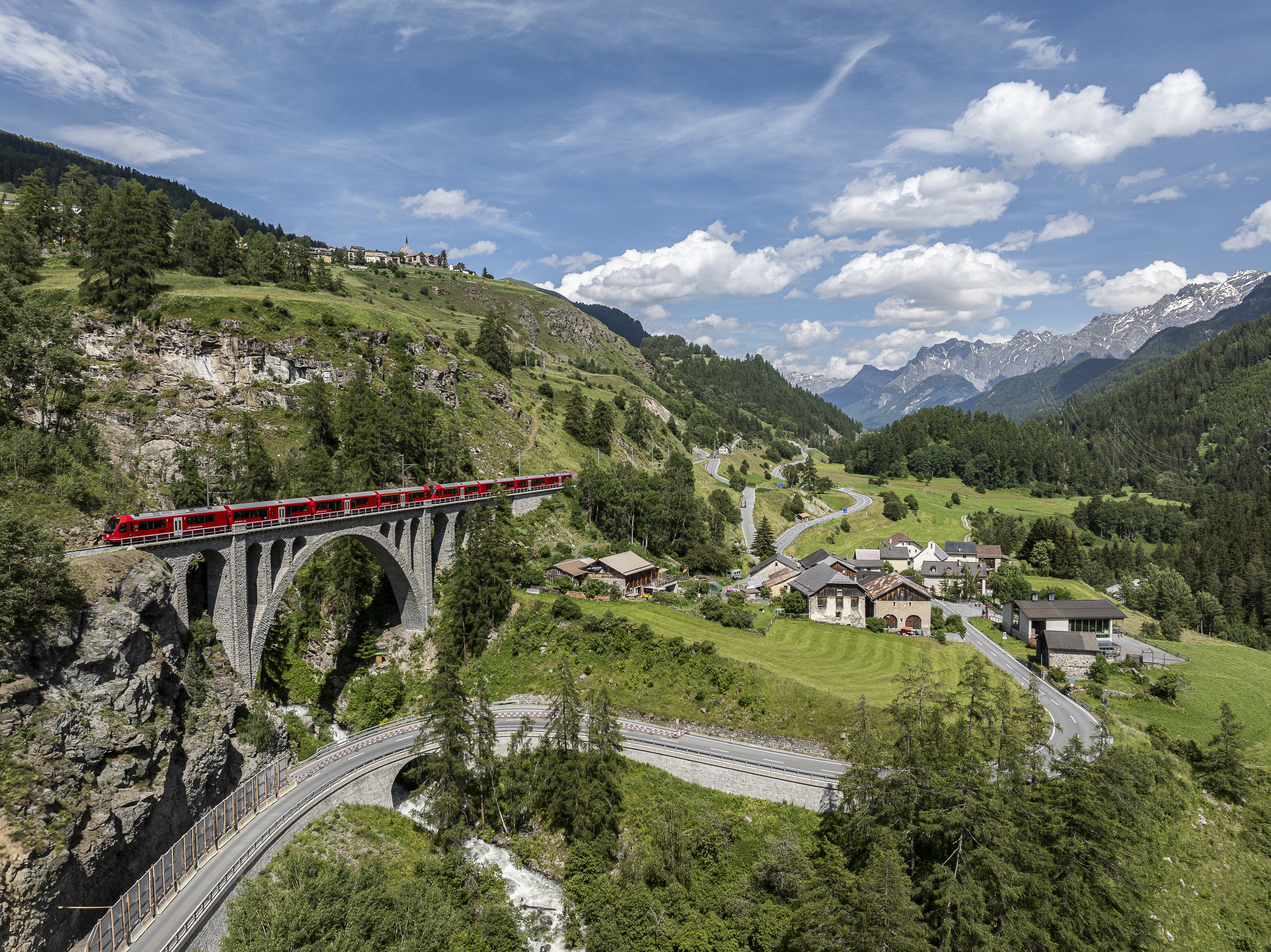Ein roter Zug überquert ein Viadukt. Darunter eine Straße durch grünes Tal. Im Hintergrund Alpen unter blauem Himmel.