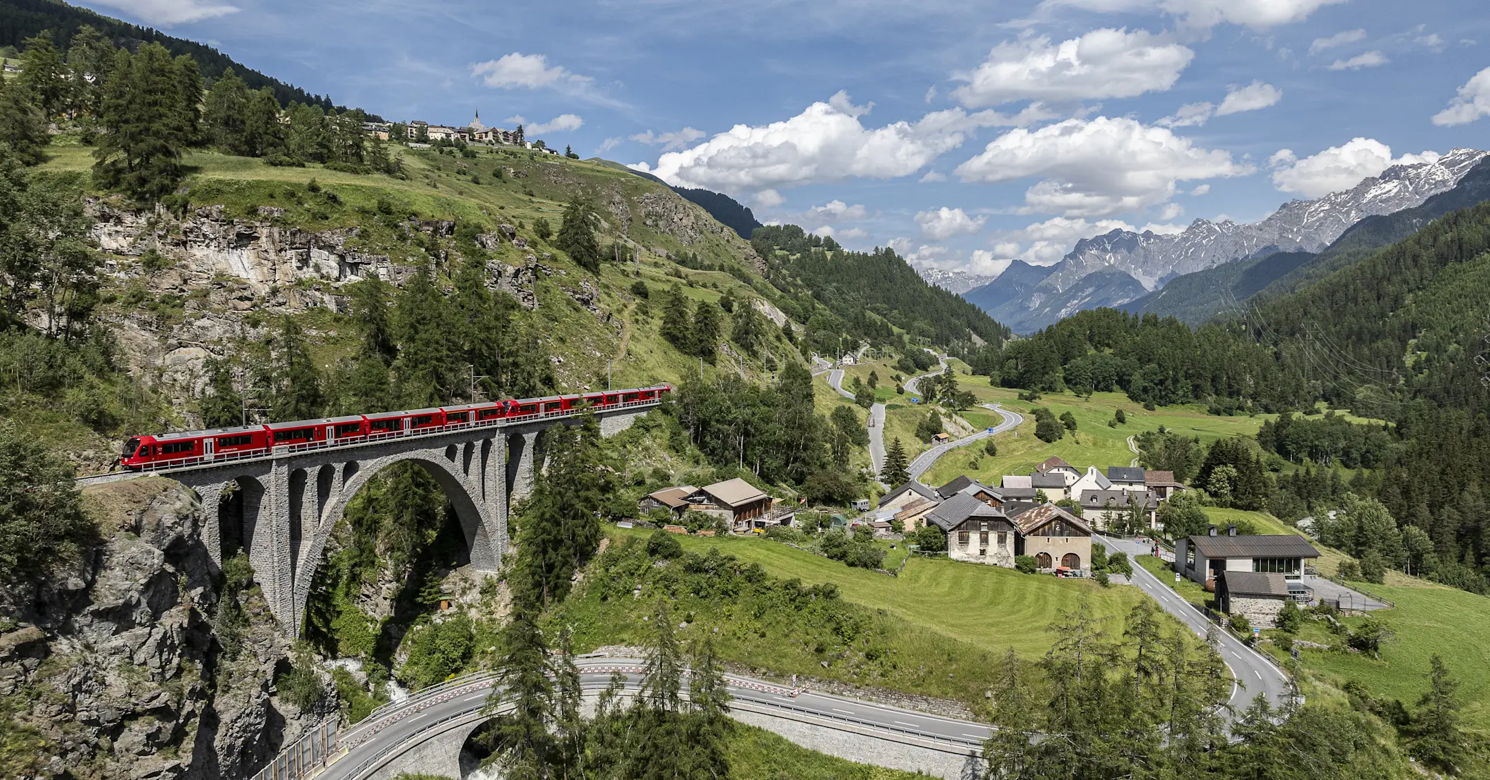 Ein roter Zug überquert ein Viadukt. Darunter eine Straße durch grünes Tal. Im Hintergrund Alpen unter blauem Himmel.