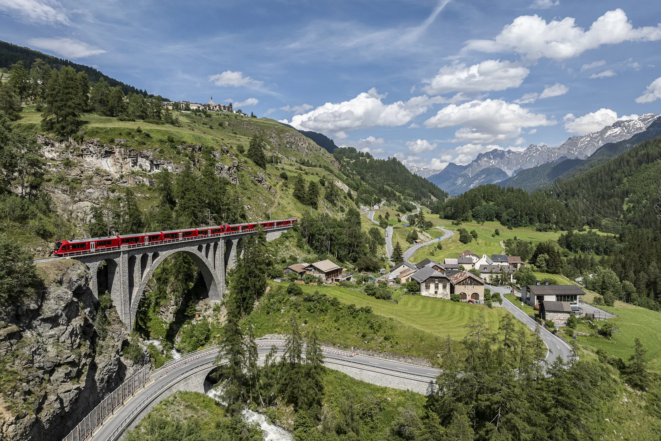 Ein roter Zug überquert ein Viadukt. Darunter eine Straße durch grünes Tal. Im Hintergrund Alpen unter blauem Himmel.