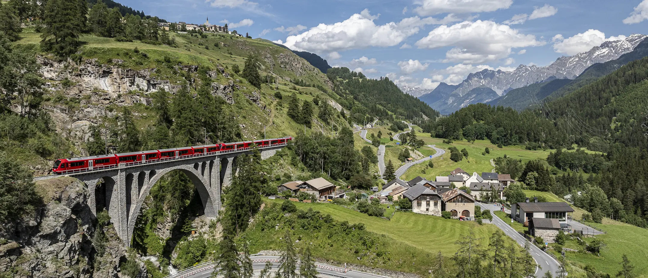 Ein roter Zug überquert ein Viadukt. Darunter eine Straße durch grünes Tal. Im Hintergrund Alpen unter blauem Himmel.