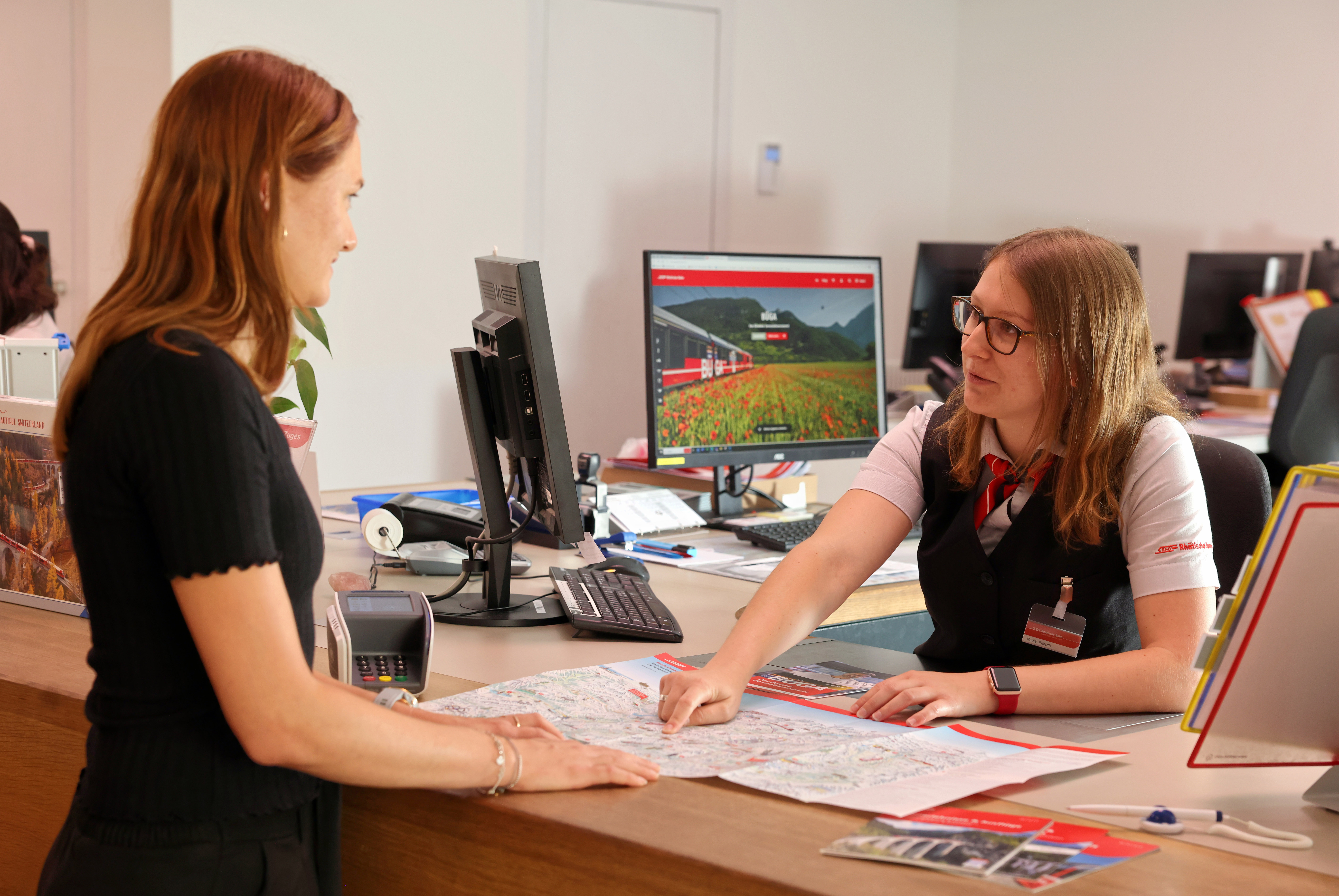 A Rhaetian Railway employee advises a customer at the service desk and points to a site plan.