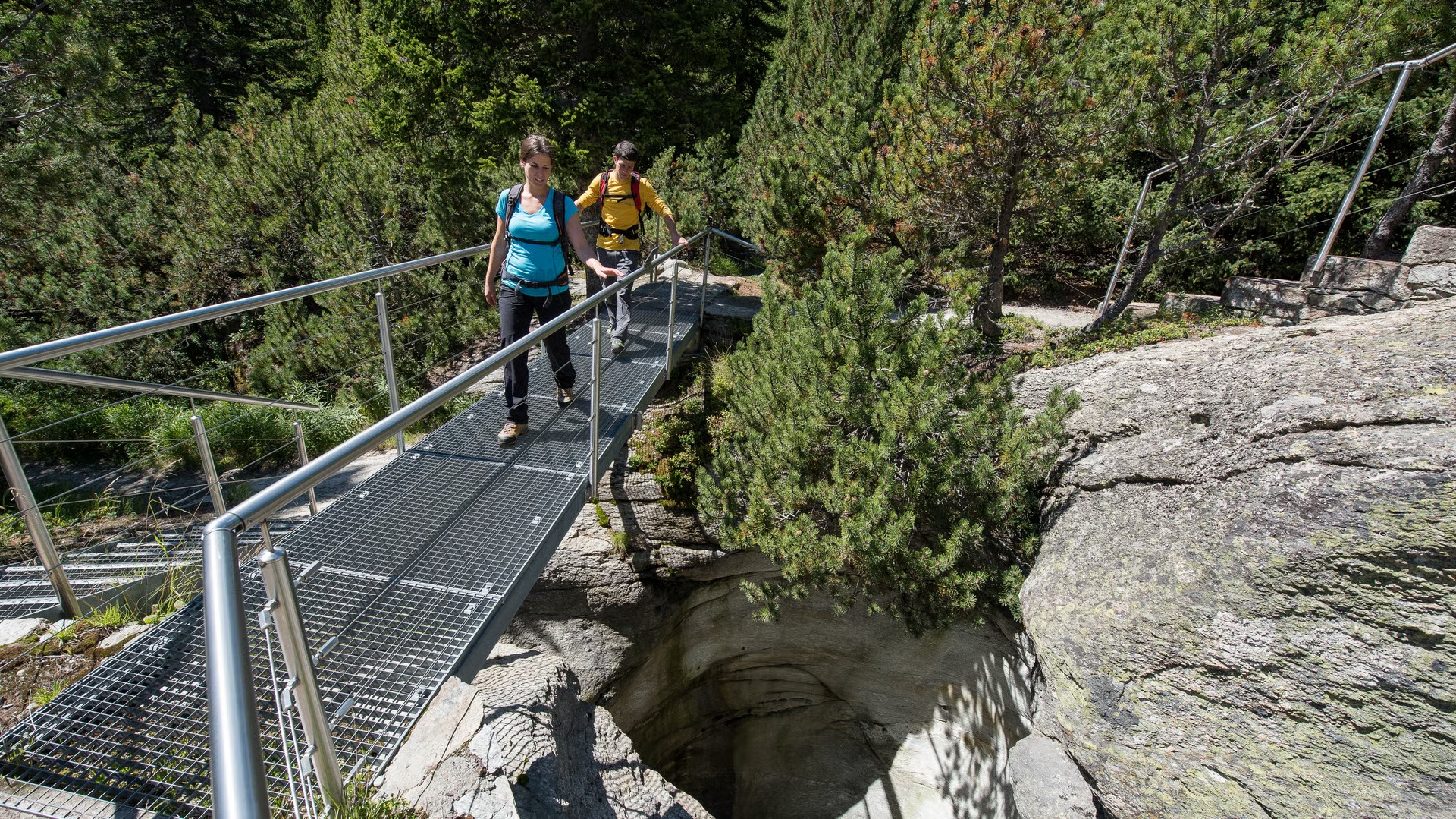 Zwei Wandernde überqueren auf einer Metallbrücke eine Gletschermühle im Gletschergarten Cavaglia, umgeben von alpinem Wald.