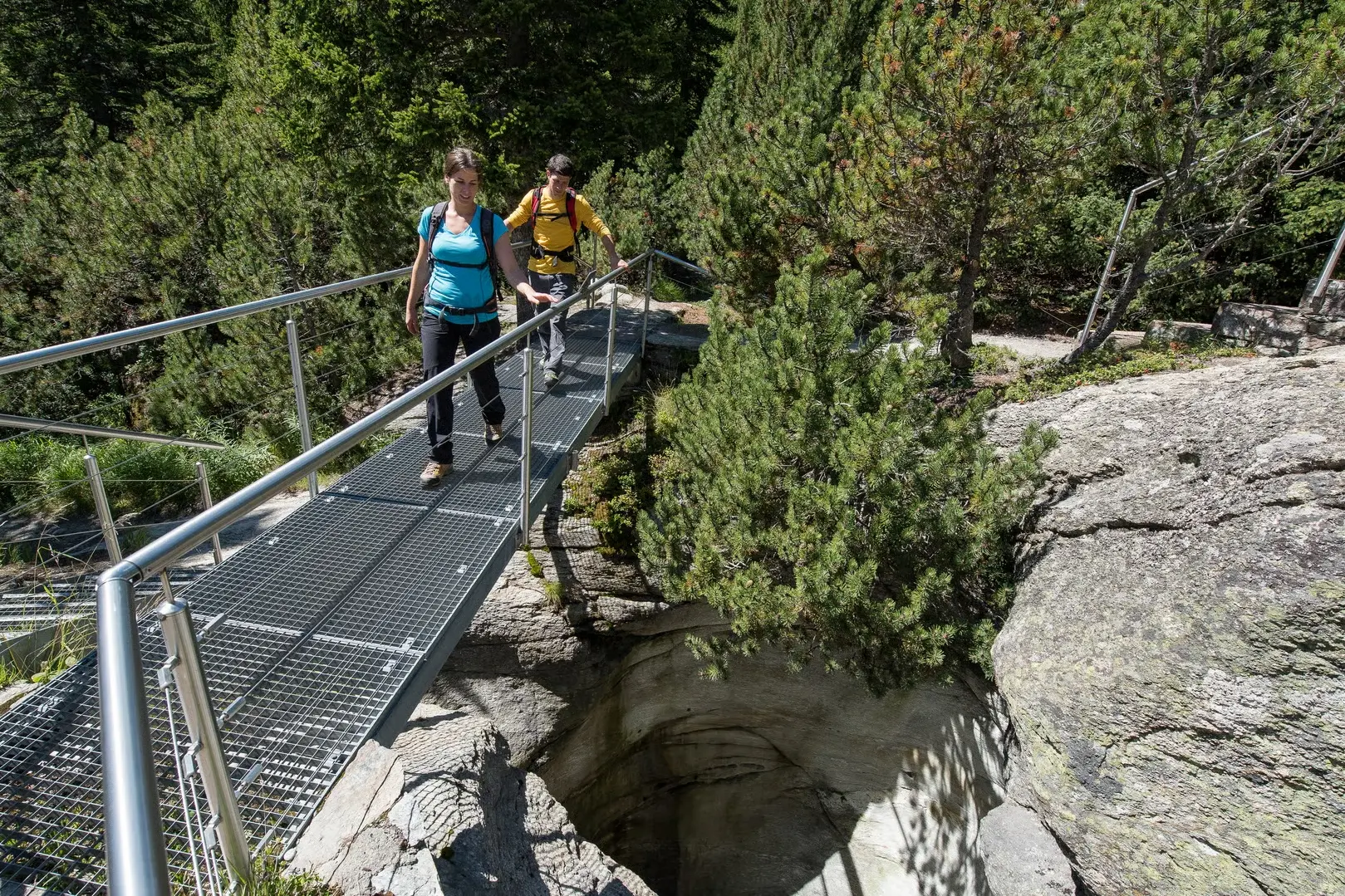 Zwei Wandernde überqueren auf einer Metallbrücke eine Gletschermühle im Gletschergarten Cavaglia, umgeben von alpinem Wald.