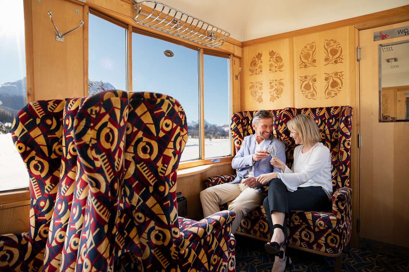 Ein Paar stösst mit Champagnergläsern in einem nostalgisch gestalteten Zugabteil des Alpine Classic Pullman an.