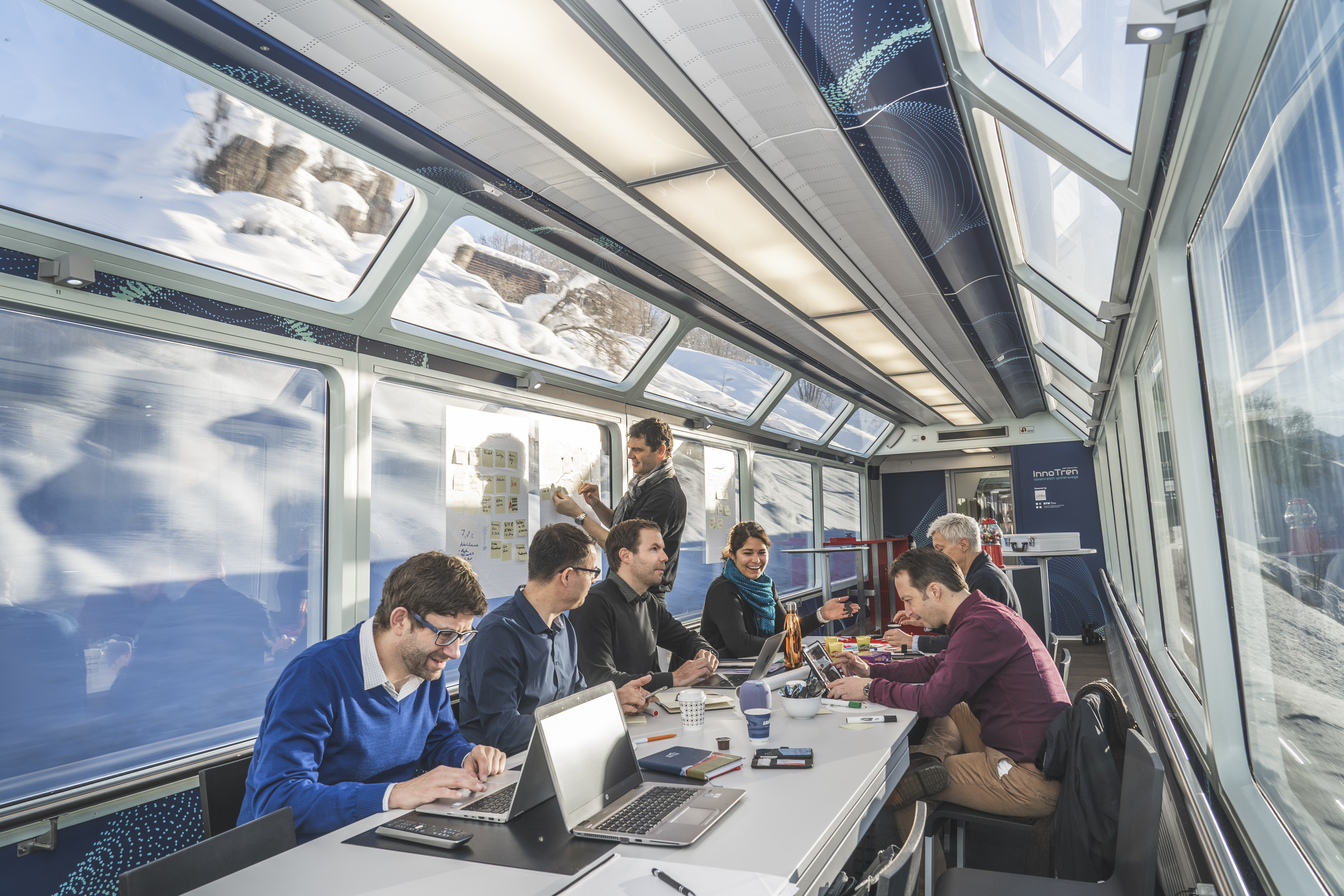 Mehrere Personen arbeiten gemeinsam an Laptops und Flipcharts in einem modernen Panoramawagen des InnoTren.