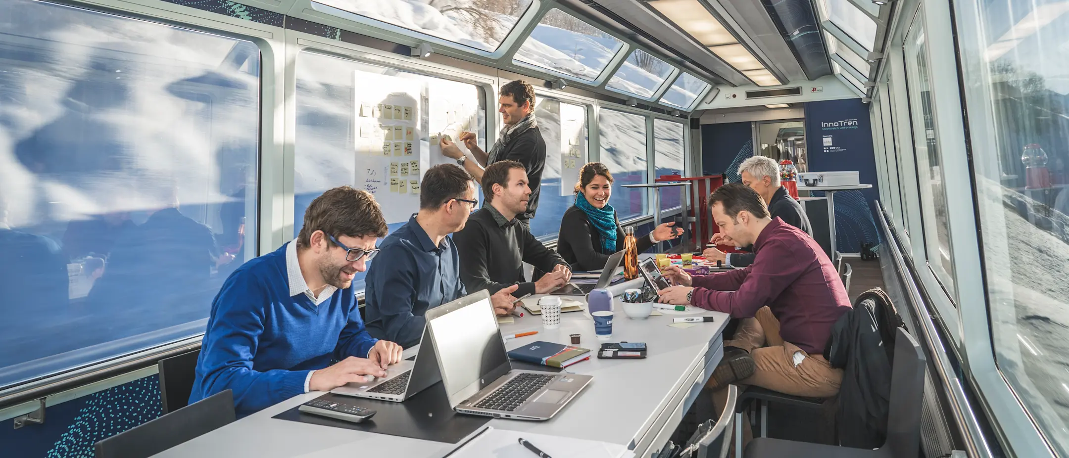 Mehrere Personen arbeiten gemeinsam an Laptops und Flipcharts in einem modernen Panoramawagen des InnoTren.