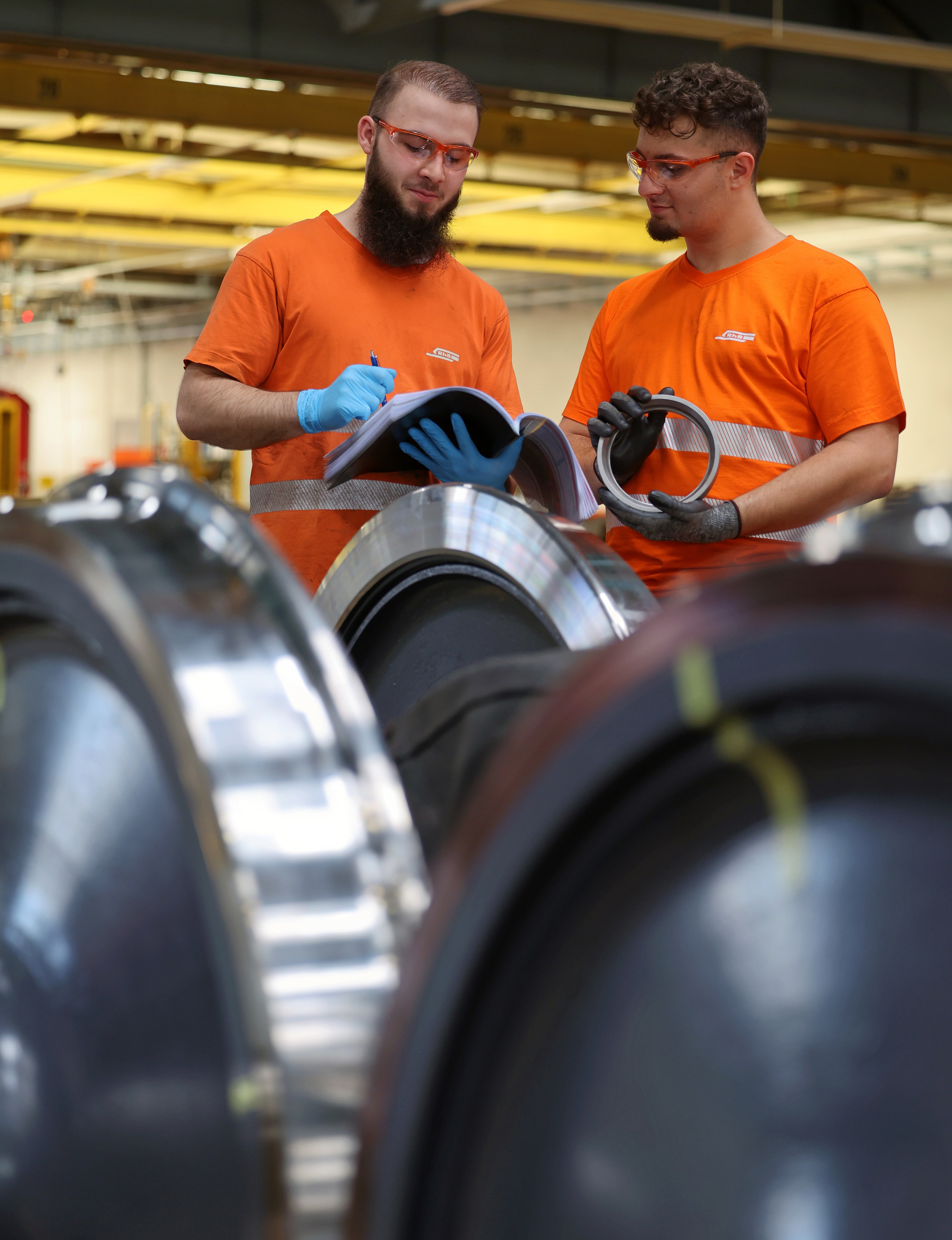 Two RhB employees in orange work clothes check wheel sets and hold technical documents in their hands.