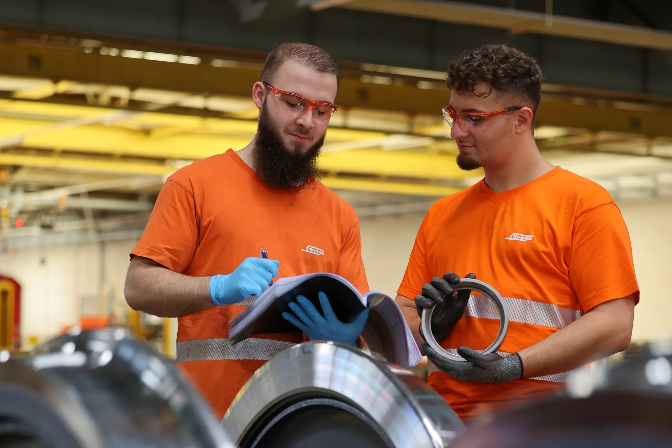 Zwei RhB-Mitarbeitende in oranger Arbeitskleidung prüfen Radsätze und halten technische Dokumente in der Hand.