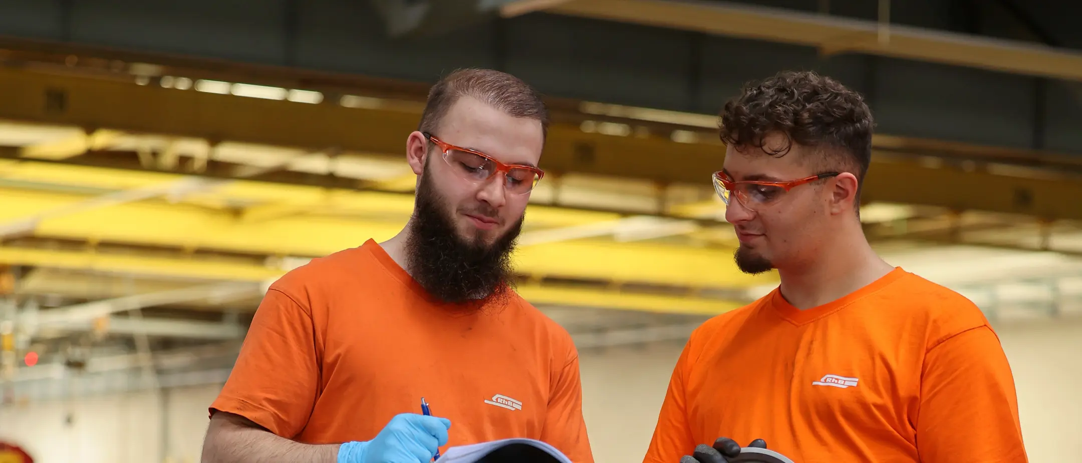 Zwei RhB-Mitarbeitende in oranger Arbeitskleidung prüfen Radsätze und halten technische Dokumente in der Hand.