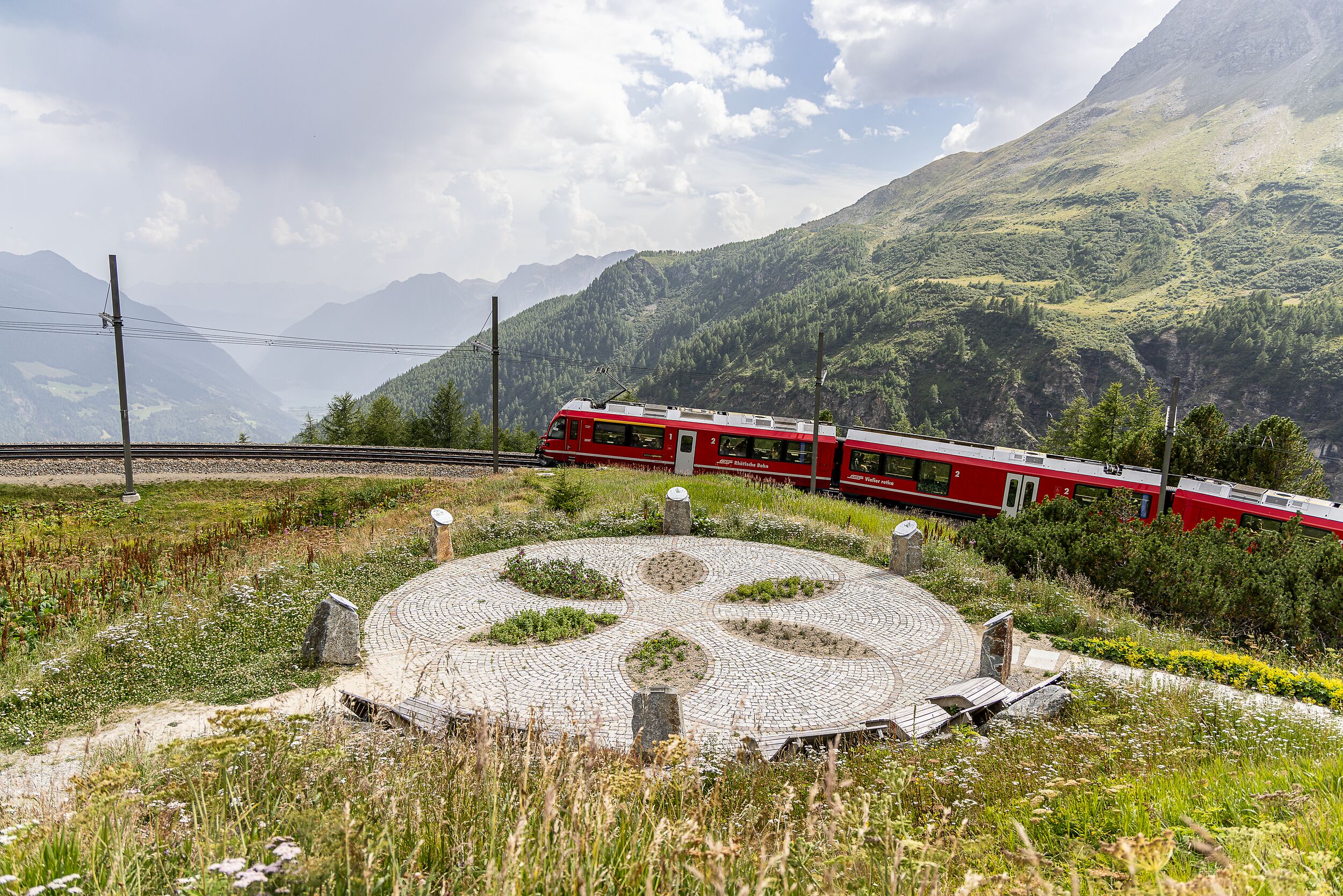 Ein roter RhB-Zug fährt durch die grüne Berglandschaft bei Alp Grüm mit Ausblick auf Tal und Gipfel.