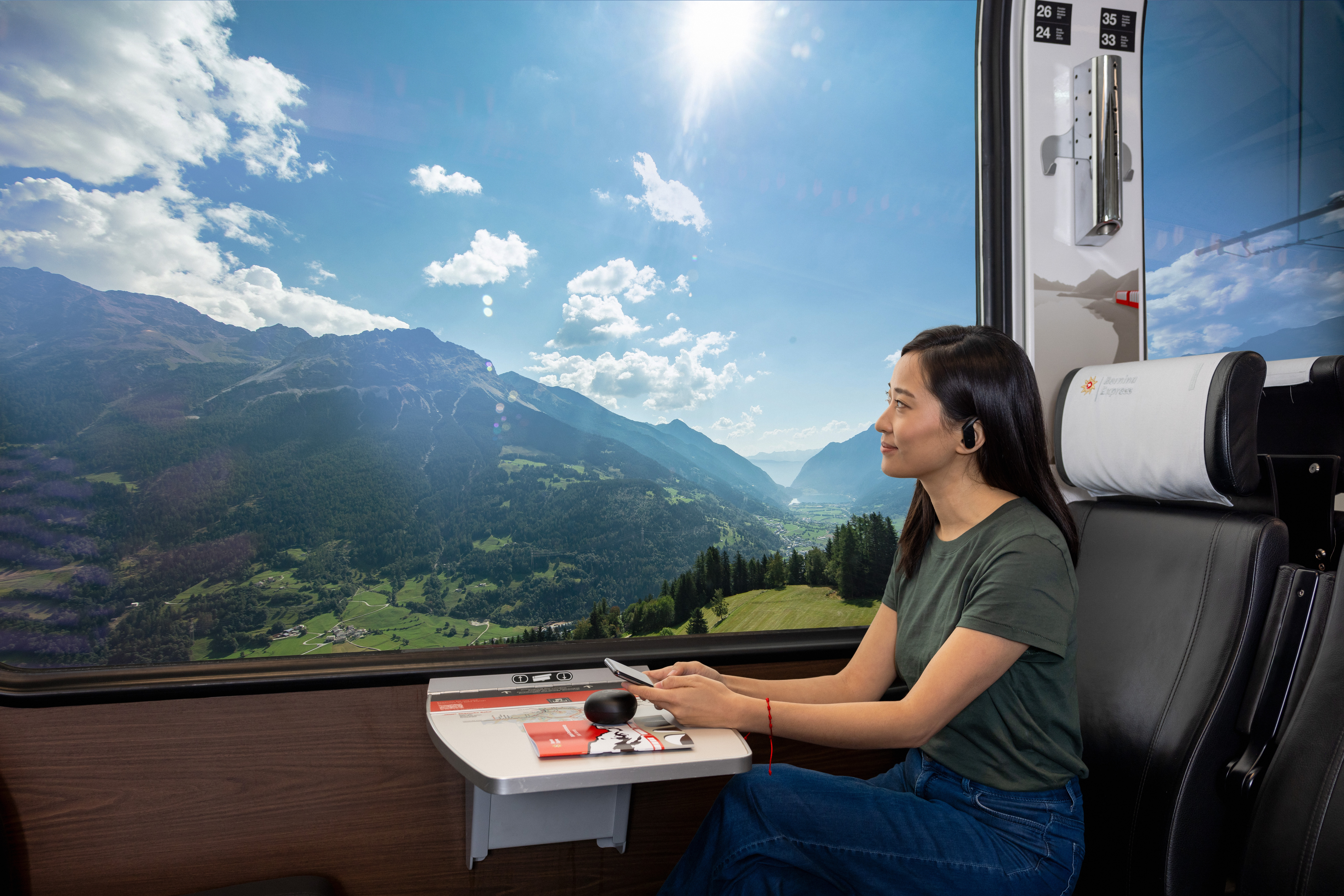 Woman sits by the window on the Bernina Express and enjoys the view of the countryside while on her mobile phone.