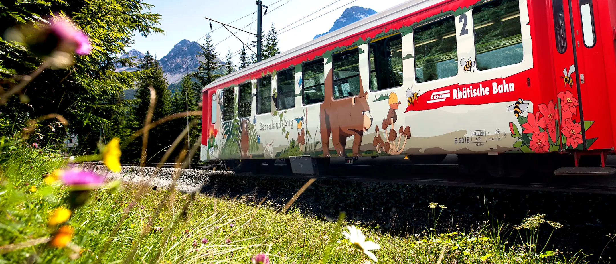 Ein bunt bemalter Wagen der Rhätischen Bahn mit Tiermotiven fährt durch eine sommerliche Berglandschaft mit Blumen.