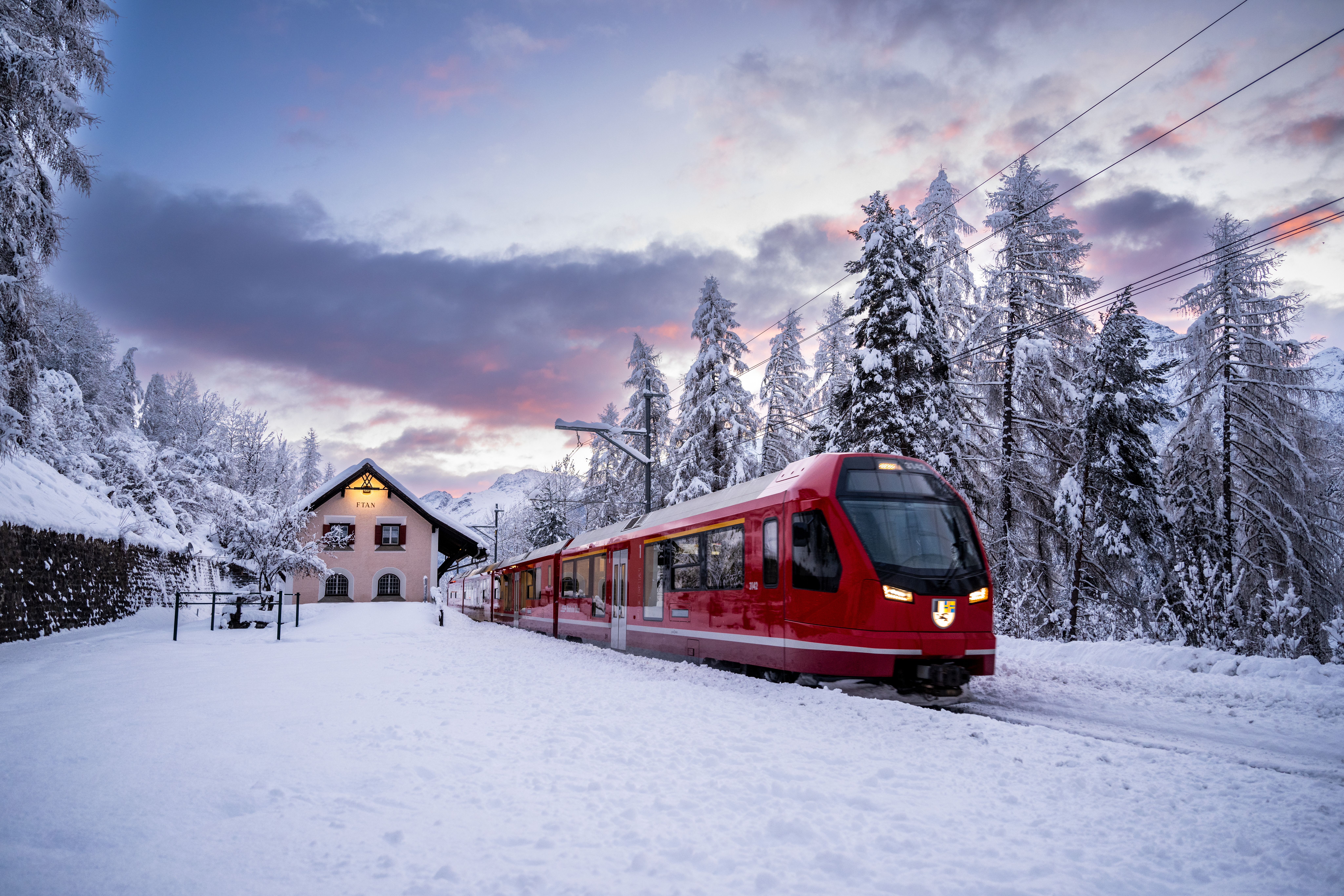 Ein Capricorn-Triebzug der Rhätischen Bahn fährt im Engadin im Winter, im Hintergrund ein Haus, schneebedeckte Landschaft