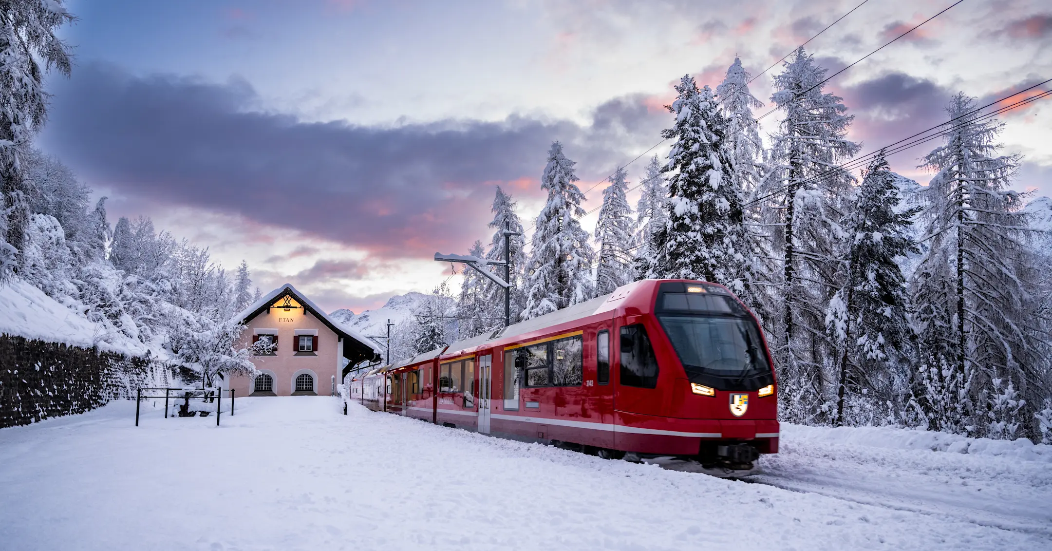 Ein Capricorn-Triebzug der Rhätischen Bahn fährt im Engadin im Winter, im Hintergrund ein Haus, schneebedeckte Landschaft