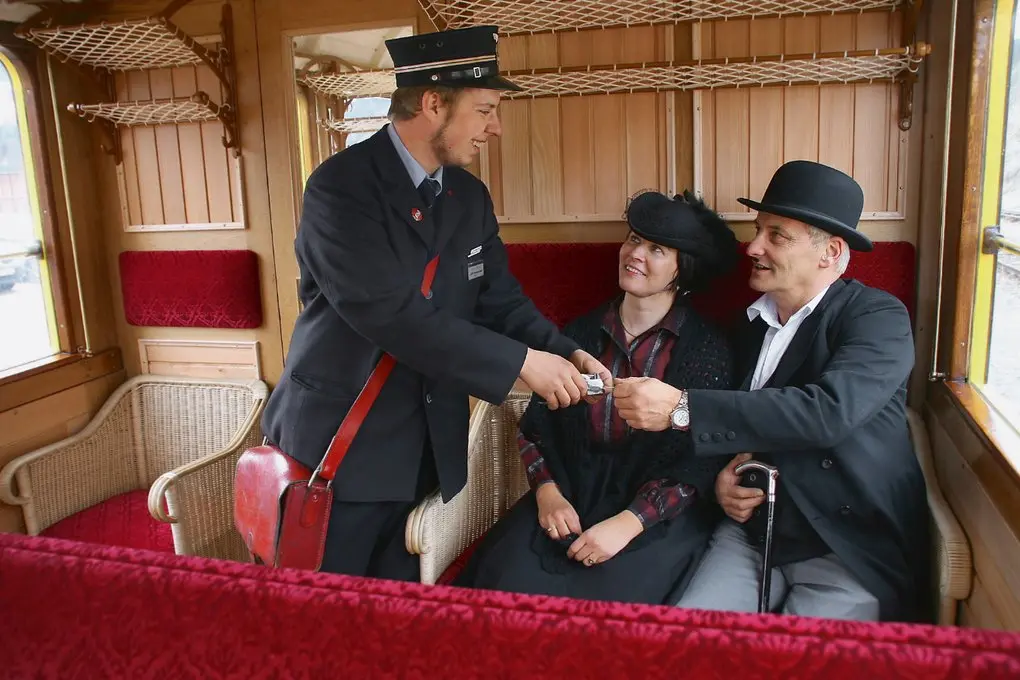 Ein Zugbegleiter in historischer Uniform kontrolliert die Fahrkarten eines Paares in einem nostalgischen Eisenbahnabteil.