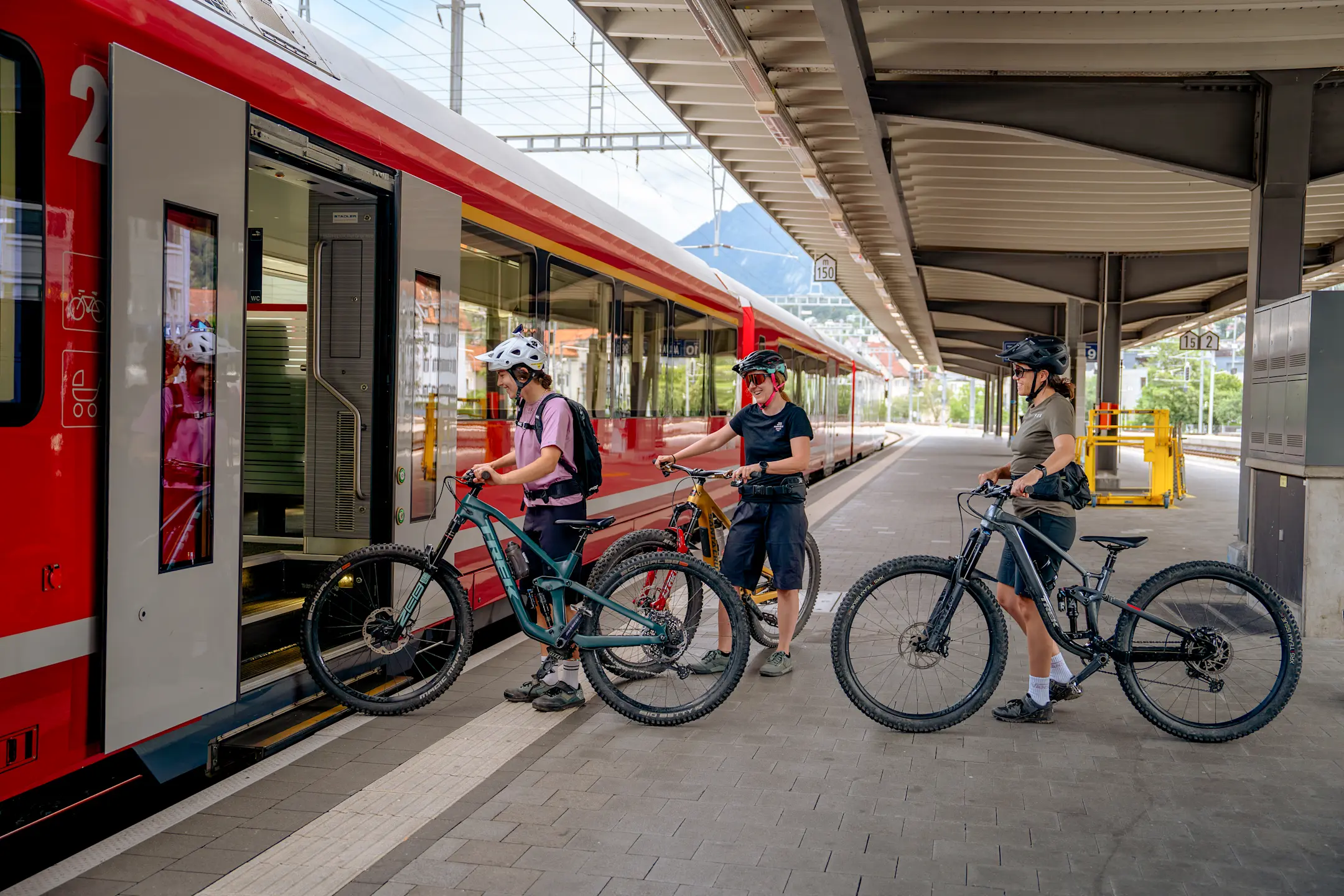 Drei Personen mit Mountainbikes steigen am Bahnhof in einen Zug mit Veloabteil ein, bereit für eine Tour durch Graubünden.