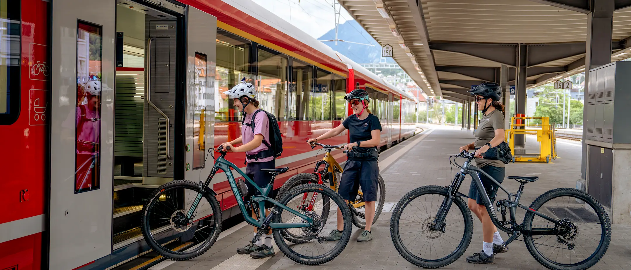 Drei Personen mit Bikes steigen am Bahnhof in einen RhB-Zug mit Veloabteil ein – bereit für eine Tour durch Graubünden.