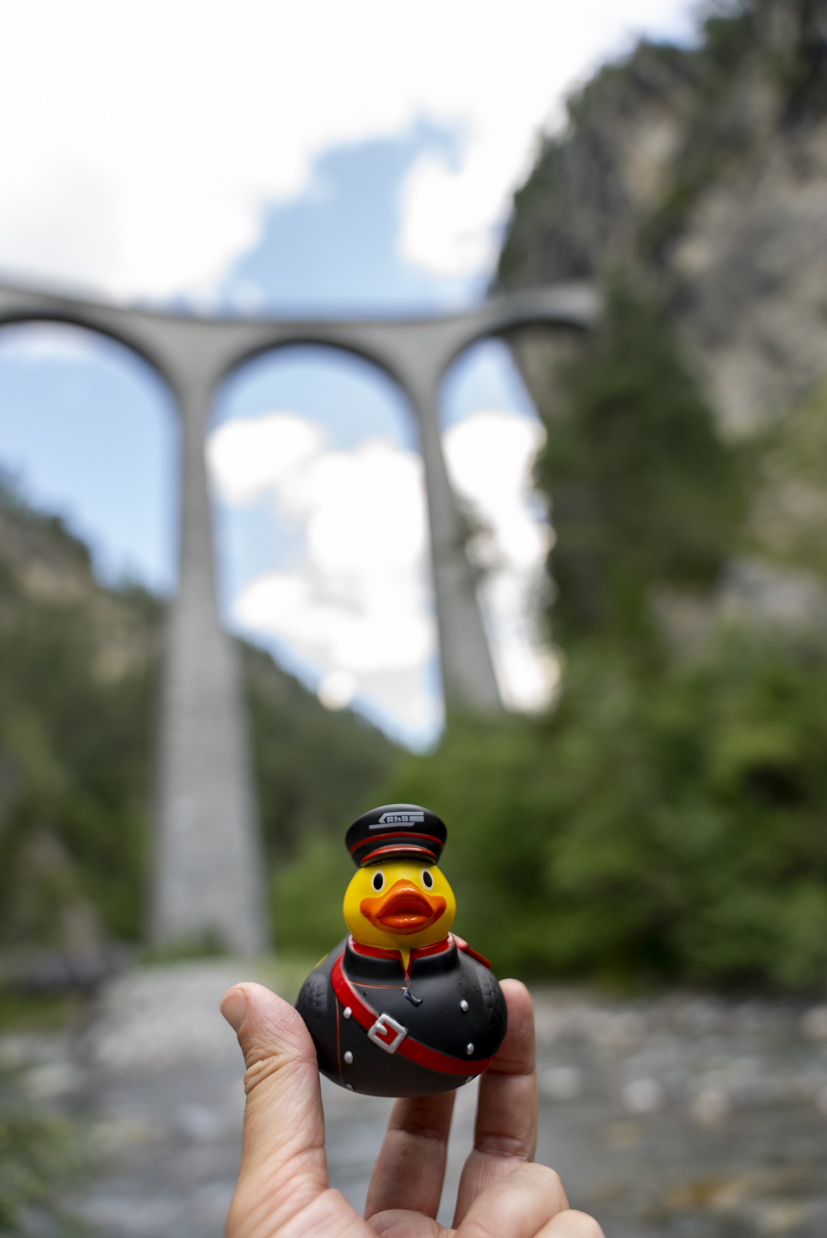 Eine gelbe Badeente wird von einer Hand hochgehalten. Im Hintergrund steht der Landwasserviadukt. 