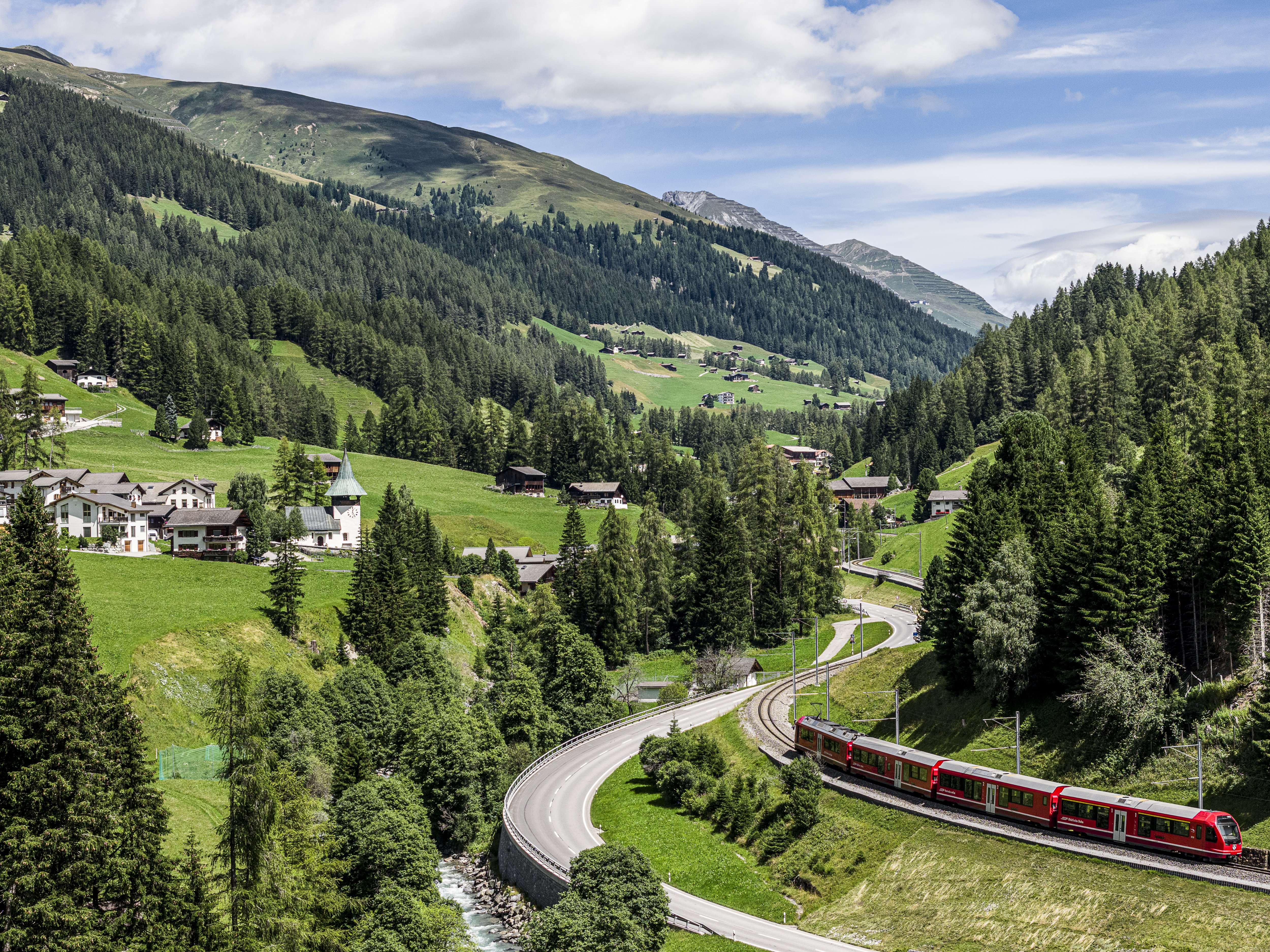 Ein roter Regiozug der RhB fährt durch das grüne Landwassertal, vorbei an Wäldern, Wiesen und typischen Walserhäusern.