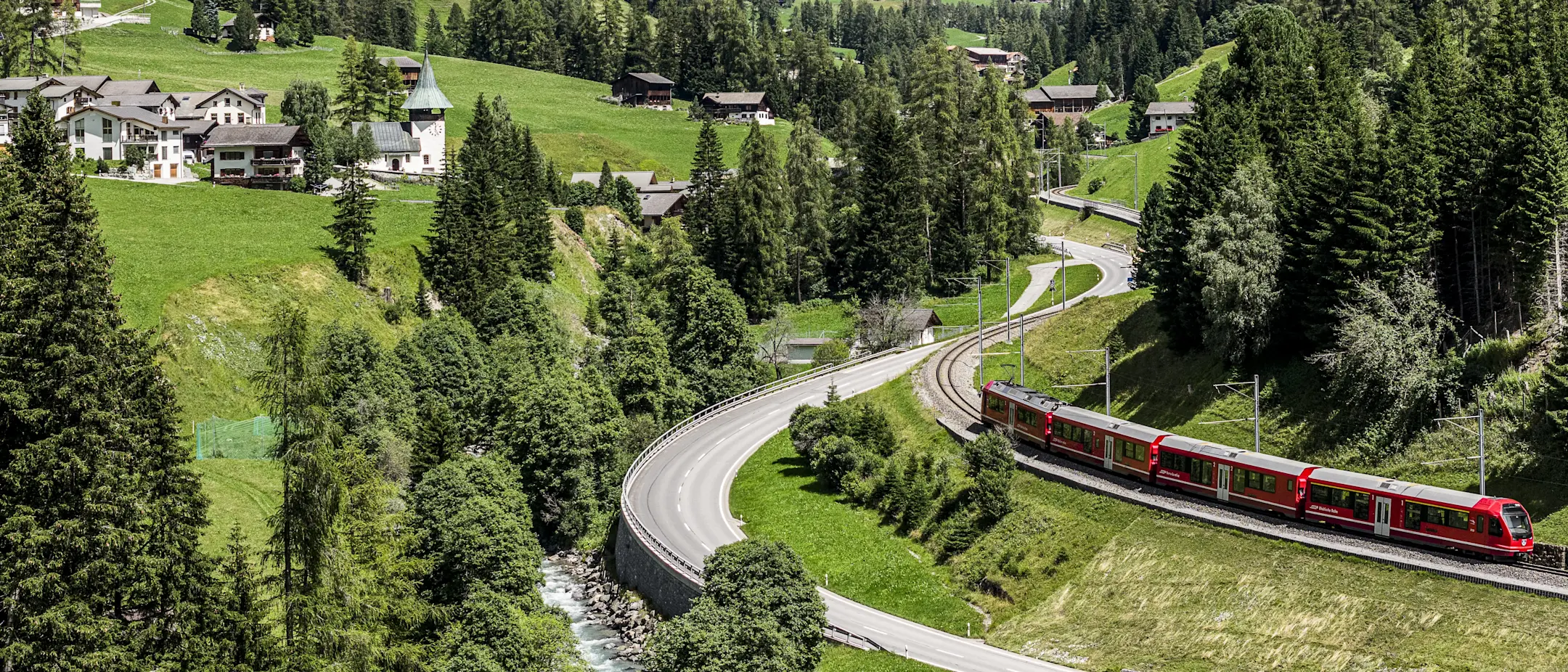 Ein roter Regiozug der RhB fährt durch das grüne Landwassertal, vorbei an Wäldern, Wiesen und typischen Walserhäusern.