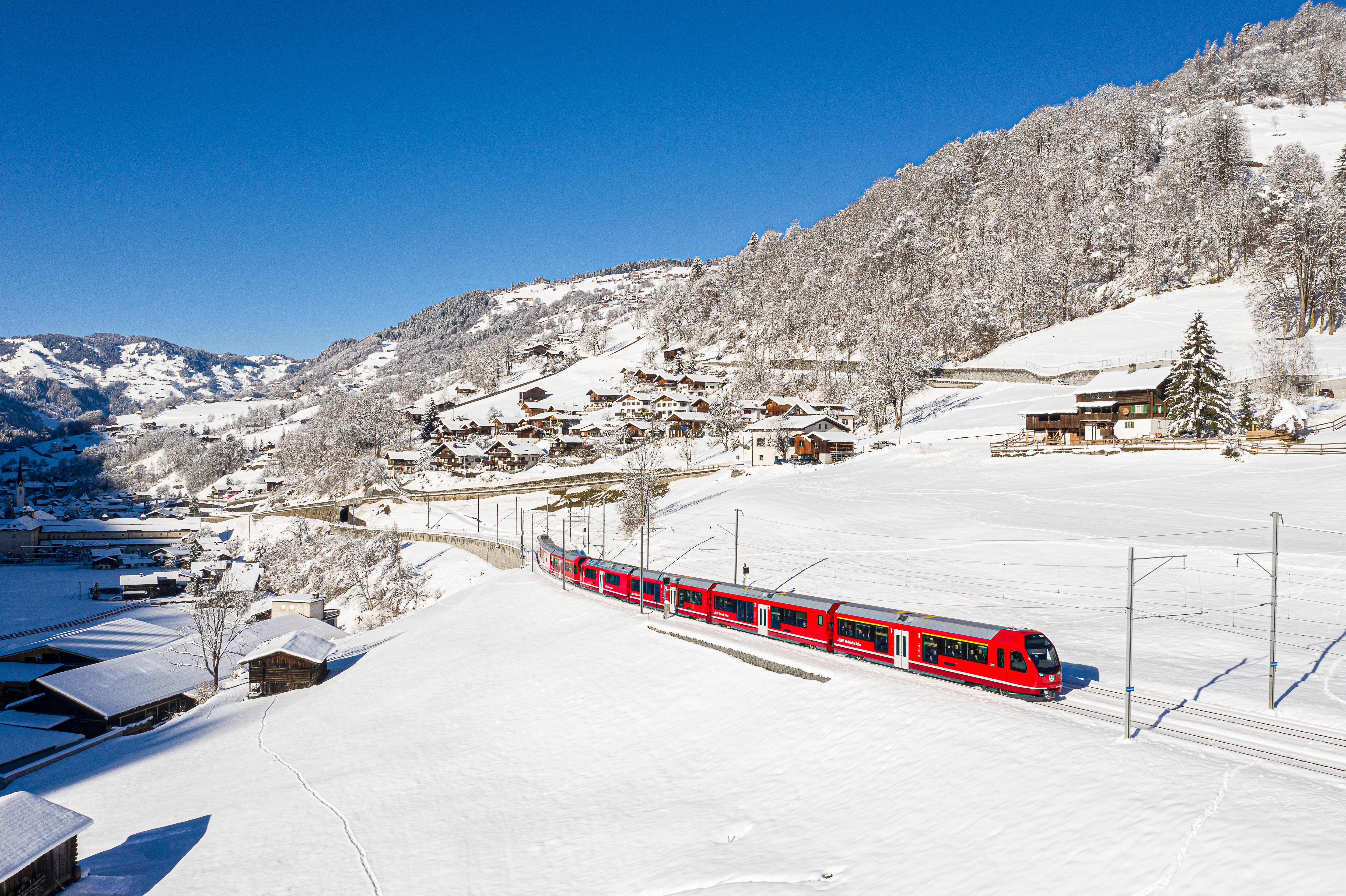 Ein Regionalzug der RhB bei Saas im Prättigau im Winter. Schneebedeckte Landschaft mit einigen Holzhäusern.