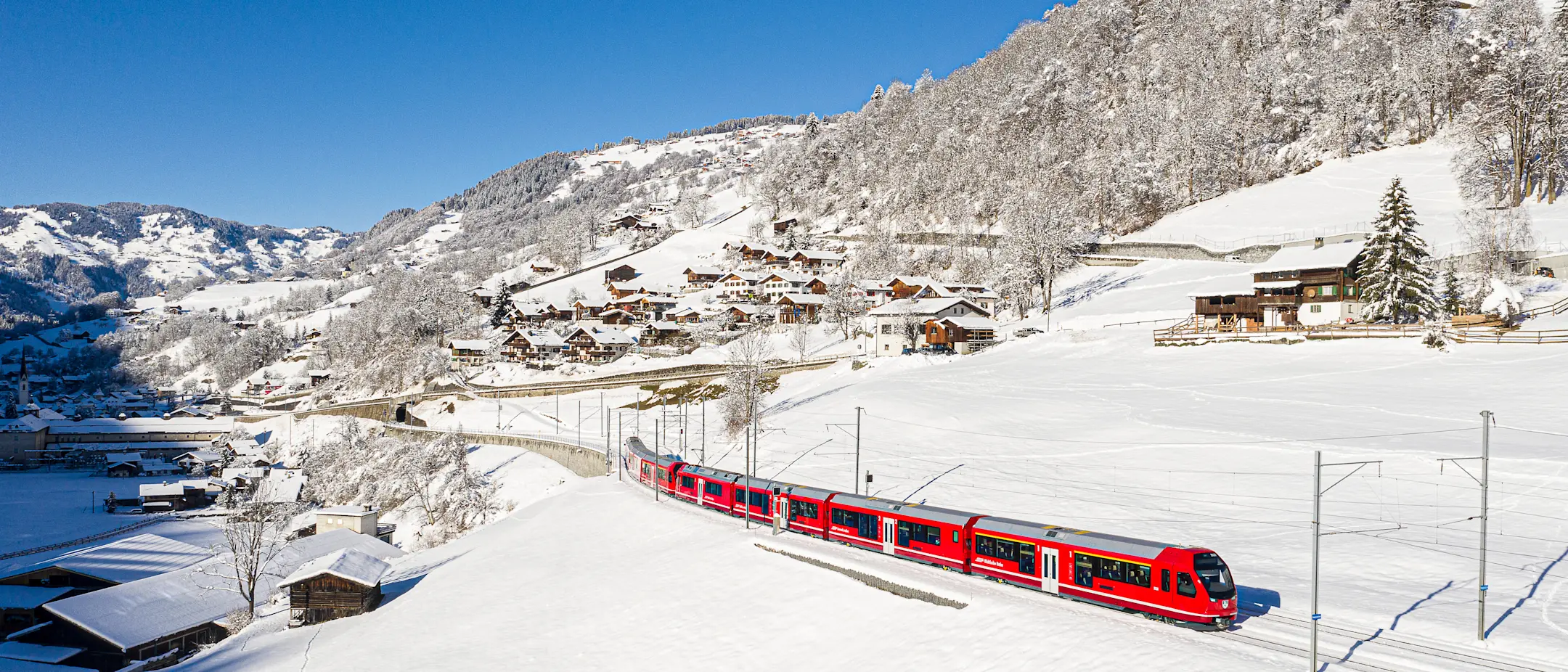 A Rhaetian Railway regional train near Saas in Prättigau in winter. Snow-covered landscape with some wooden houses.