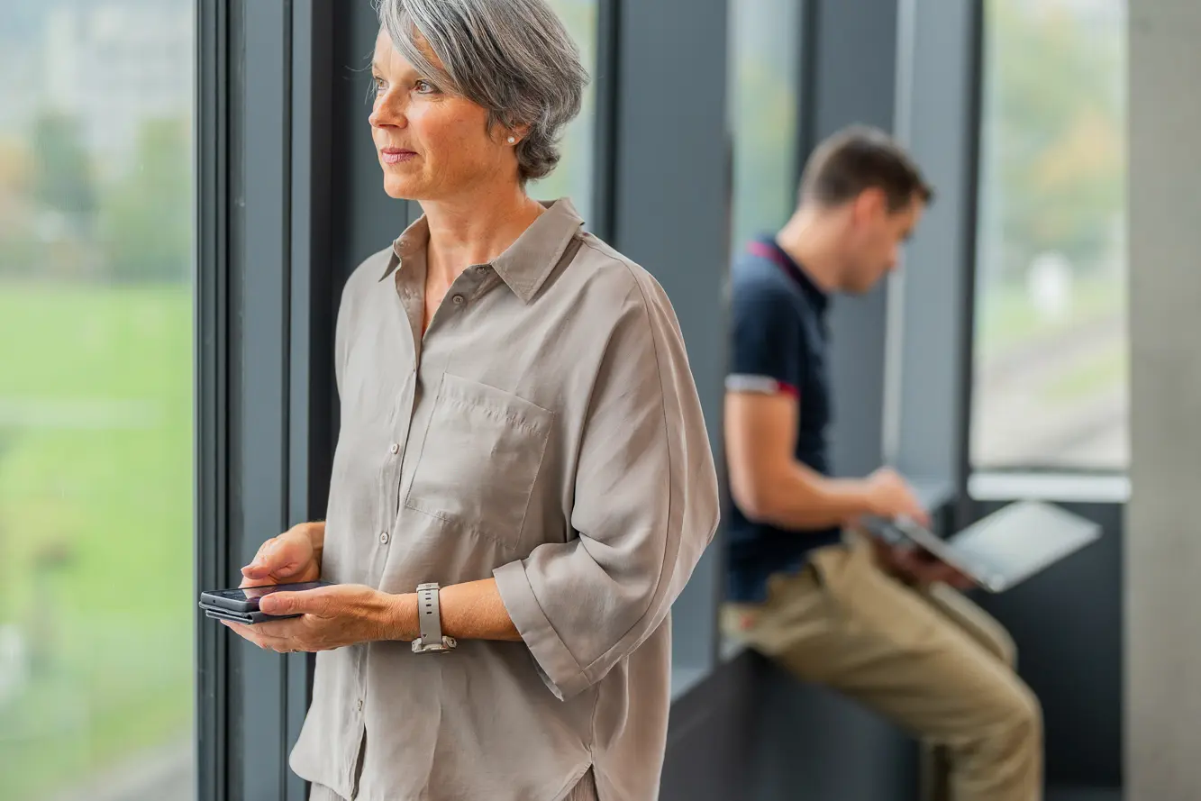 Eine Mitarbeiterin am Fenster mit Mobiltelefon; im Hintergrund ein Mann mit Notebook