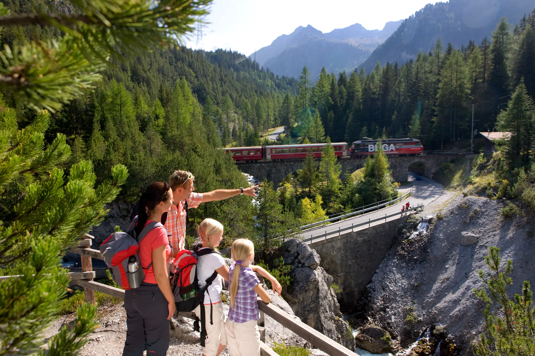 Eine Familie wandert und beobachtet einen vorbeifahrenden Zug der RhB inmitten einer alpinen Wald- und Berglandschaft.