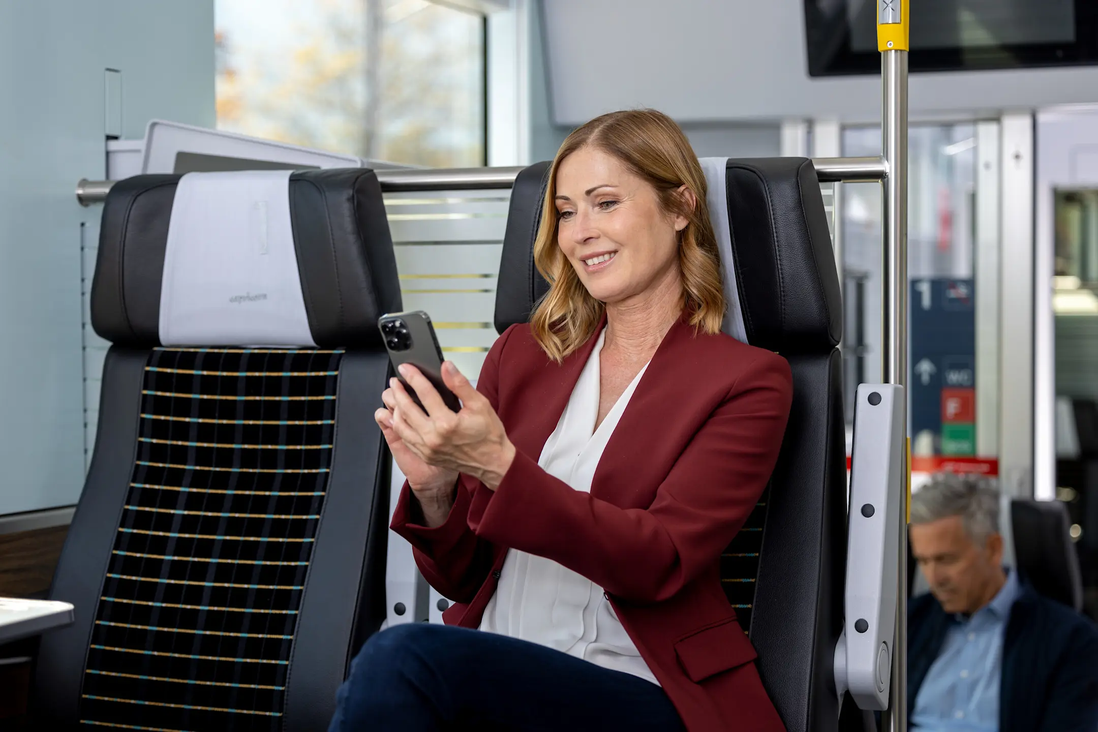 Eine Frau in einem roten Blazer sitzt in der 1. Klasse eines Capricorn-Triebzugs und schaut lächelnd auf ihr Smartphone.