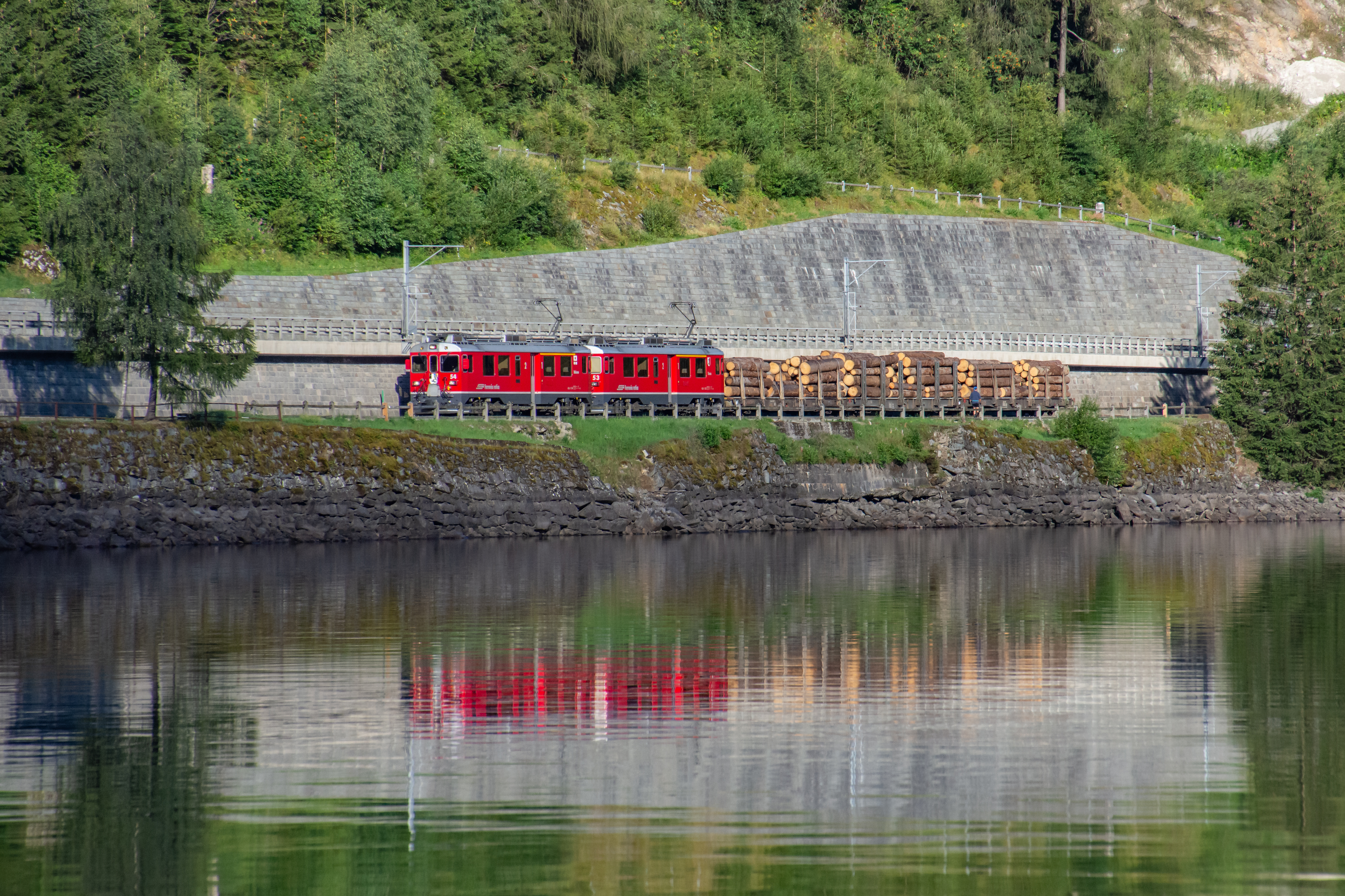 Holztransport der RhB am Lago di Poschiavo, im Vordergrund spiegelt sich der Zug im Wasser