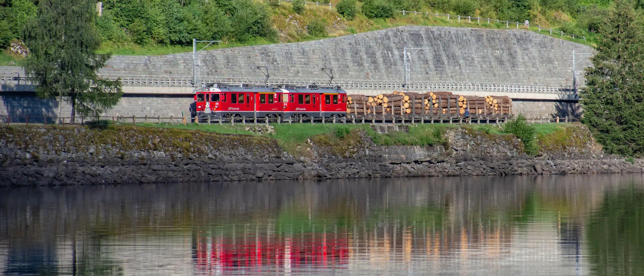 Holztransport der RhB am Lago di Poschiavo, im Vordergrund spiegelt sich der Zug im Wasser