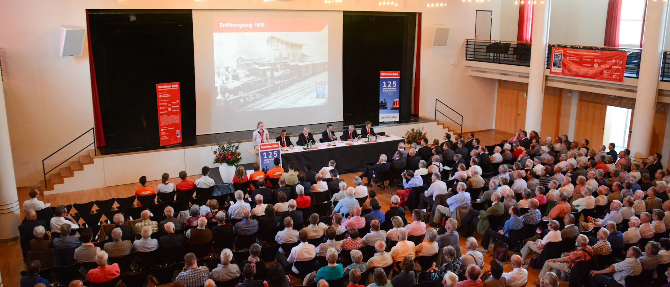 Blick von der Empore im Forum Ried hinunter auf die Teilnehmenden der Generalversammlung der RhB mit Bühne und Leinwand.