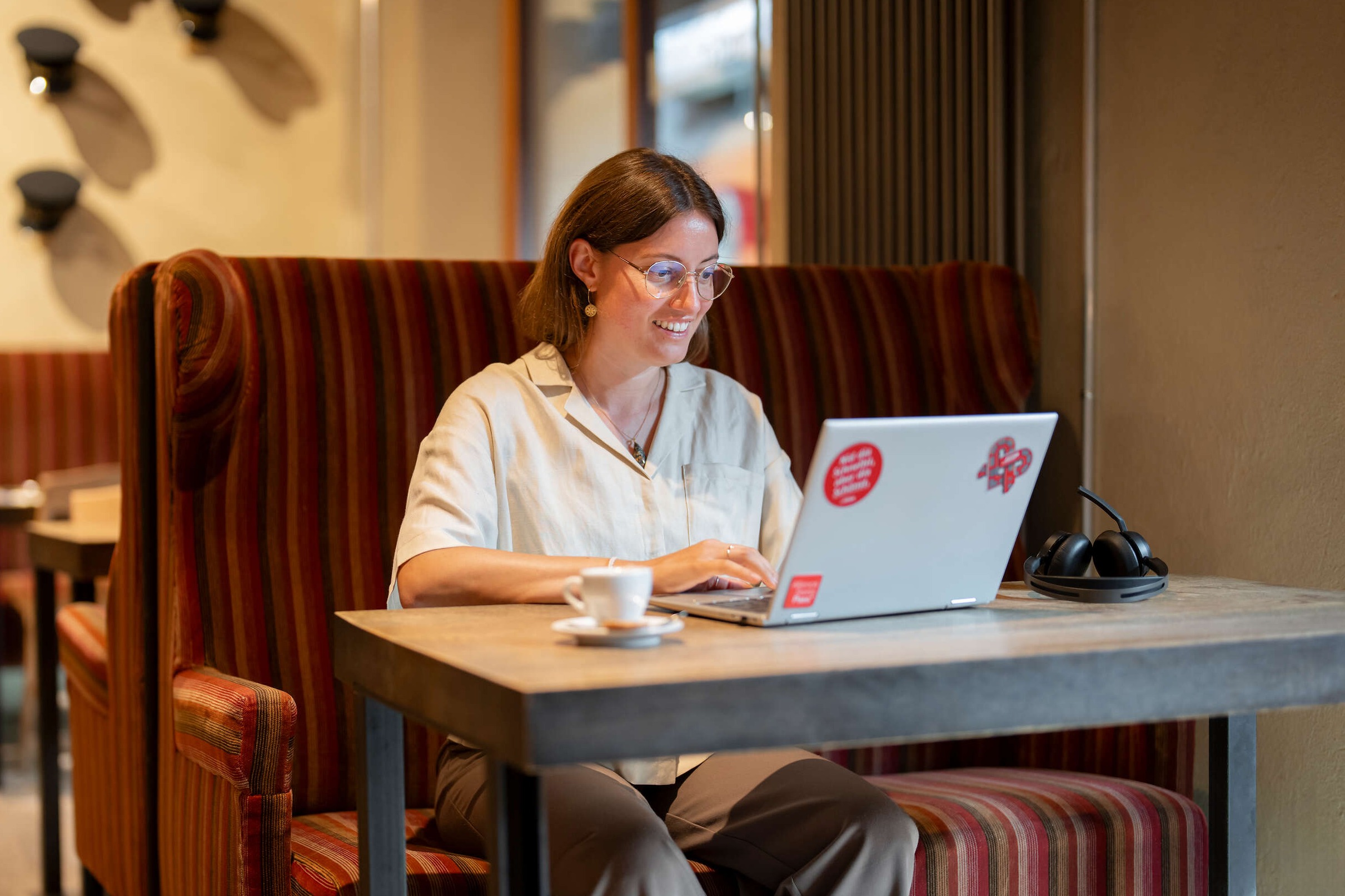 Person nimmt in einem Café an einem Laptop eine Buchung vor; auf dem Tisch stehen eine Kaffeetasse und Kopfhörer.