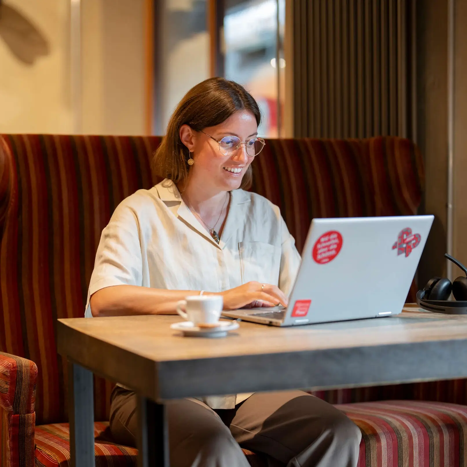 Person nimmt in einem Café an einem Laptop eine Buchung vor; auf dem Tisch stehen eine Kaffeetasse und Kopfhörer.