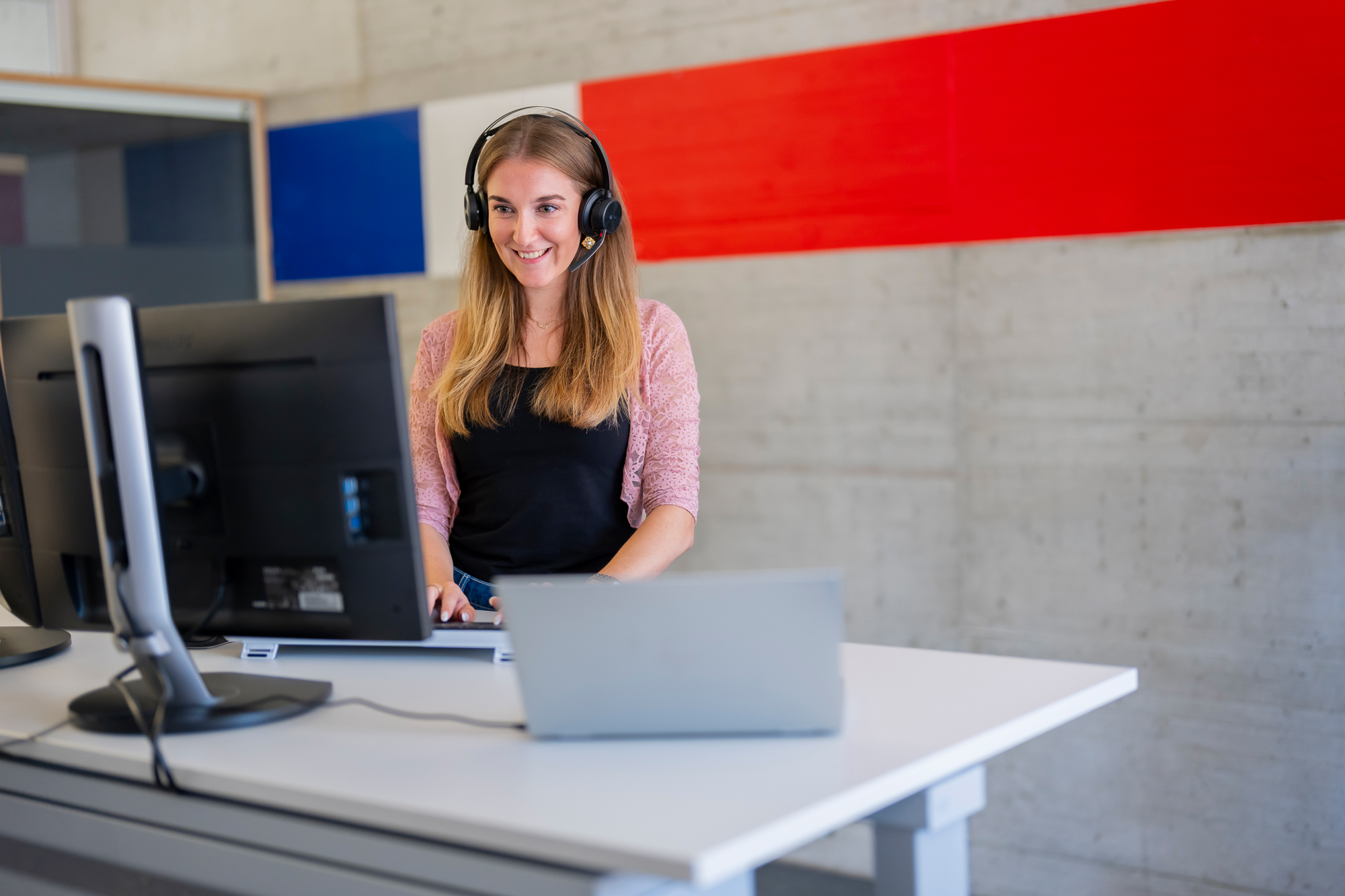 An employee works on a PC and laptop with headphones.