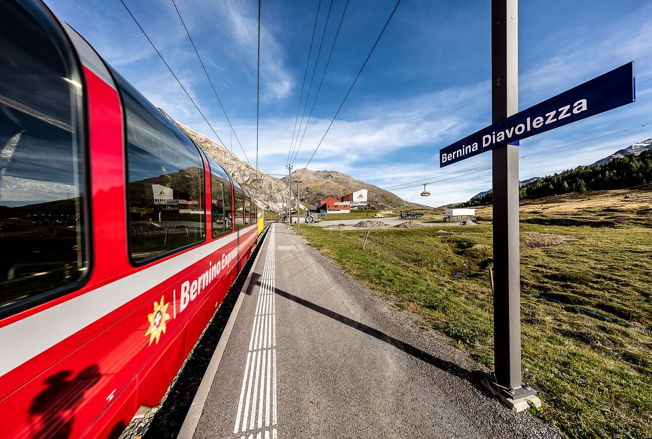Der Bernina Express hält an der Station Bernina Diavolezza inmitten einer alpinen Landschaft mit Bergen und Seilbahn.