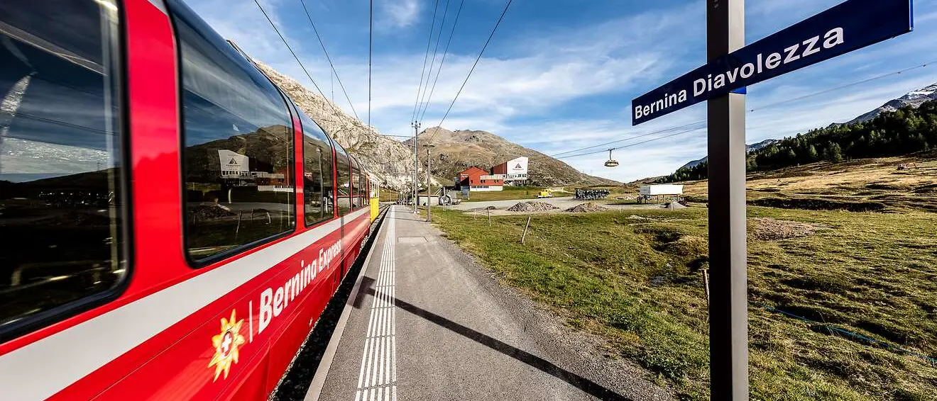 Der Bernina Express hält an der Station Bernina Diavolezza inmitten einer alpinen Landschaft mit Bergen und Seilbahn.
