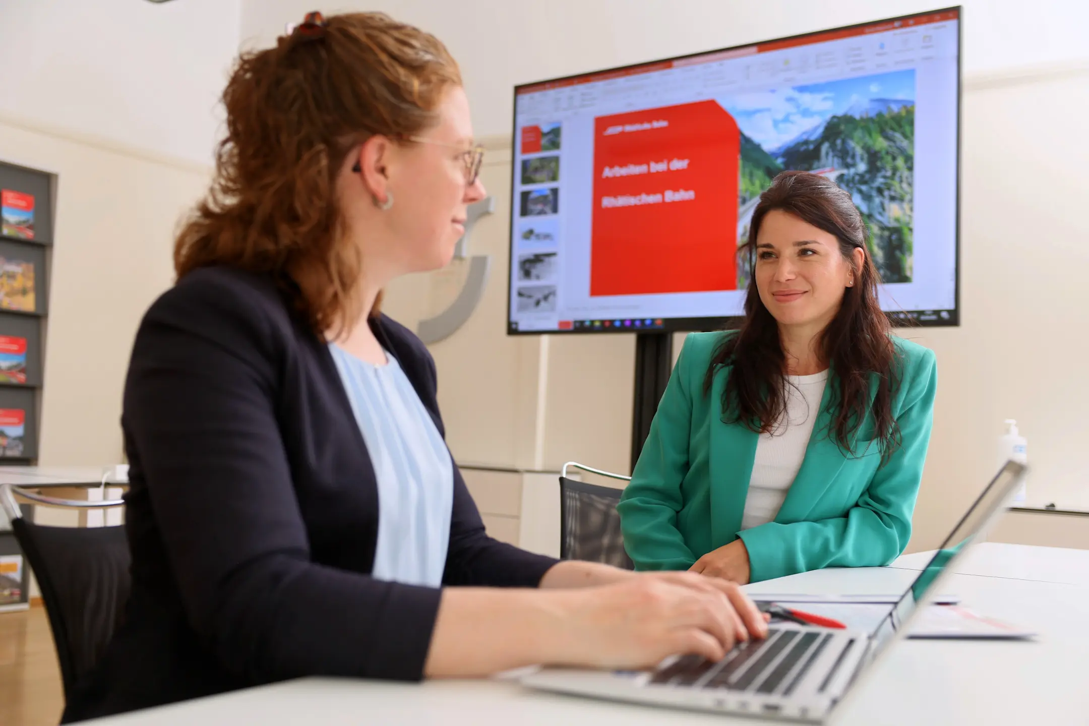 Zwei RhB-Mitarbeiterinnen in einem Büro mit Notebook und Bildschirm im Hintergrund.