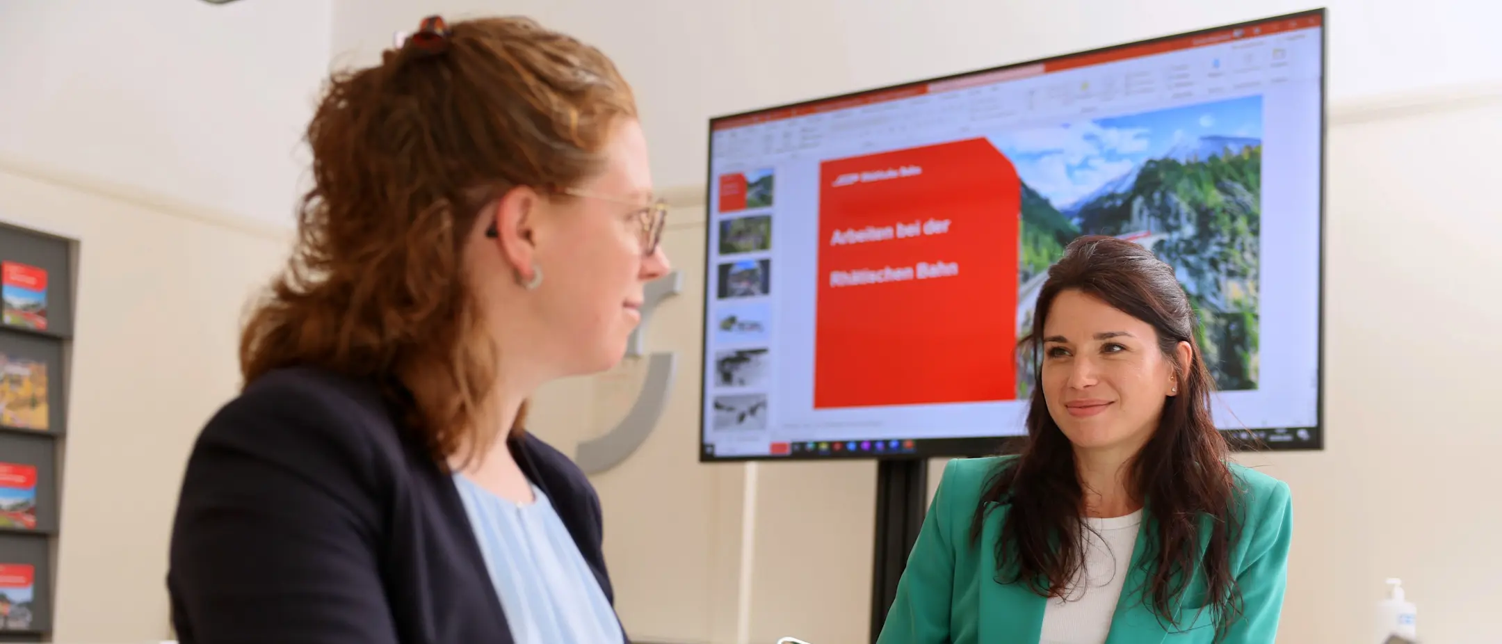 Zwei RhB-Mitarbeiterinnen in einem Büro mit Notebook und Bildschirm im Hintergrund.