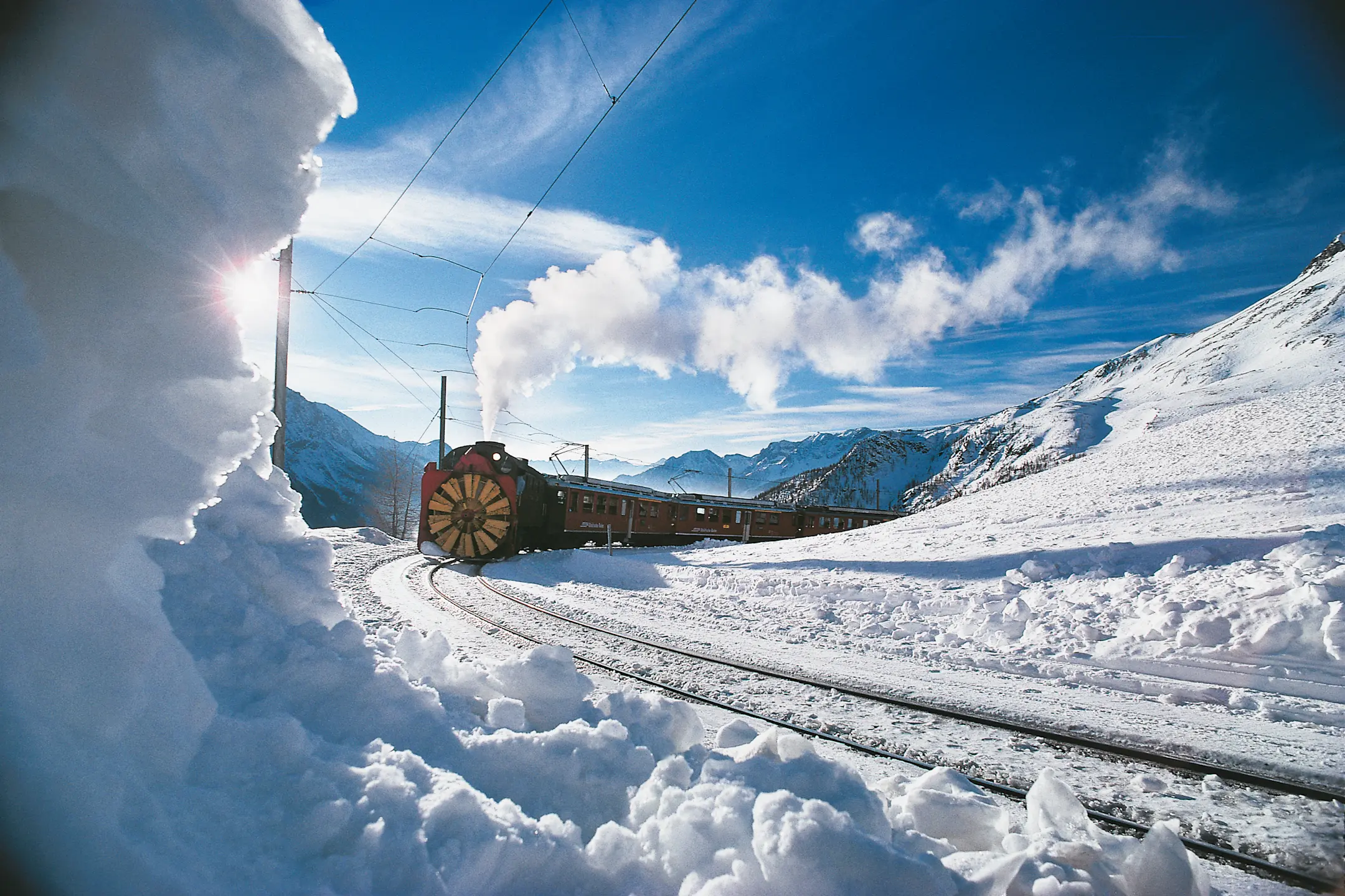 Dampfschneeschleuder der Rhätischen Bahn räumt verschneite Bahngleise auf dem Berninapass bei sonnigem Winterwetter frei.