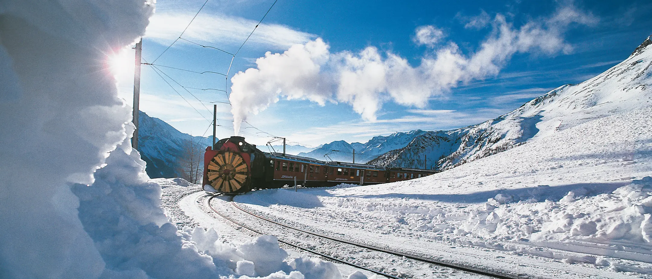 Dampfschneeschleuder der Rhätischen Bahn räumt verschneite Bahngleise auf dem Berninapass bei sonnigem Winterwetter frei.