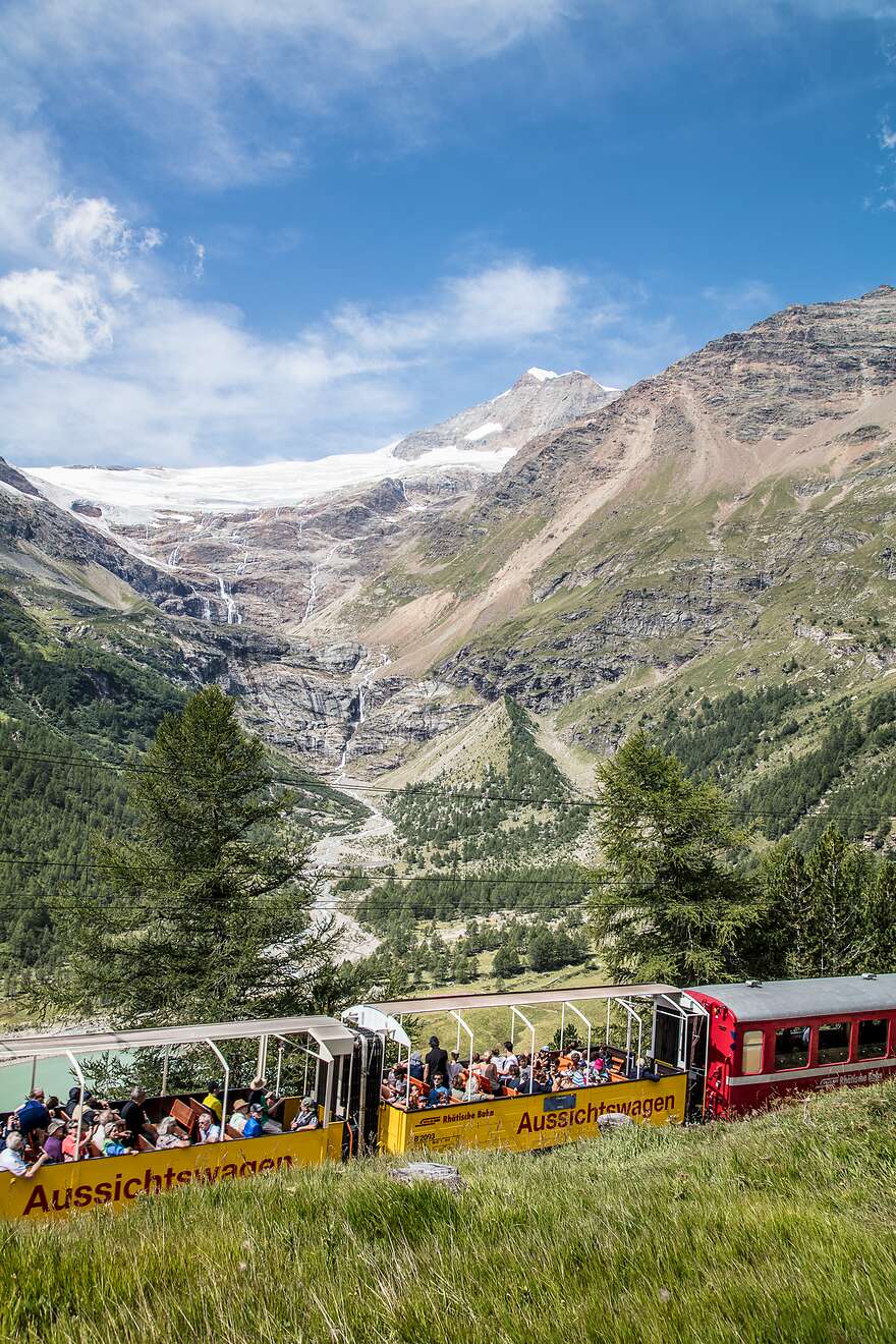 Ein gelber offener Aussichtswagen der RhB fährt durch eine grüne Berglandschaft bei Alp Grüm. Im Hintergrund ein Gletscher.
