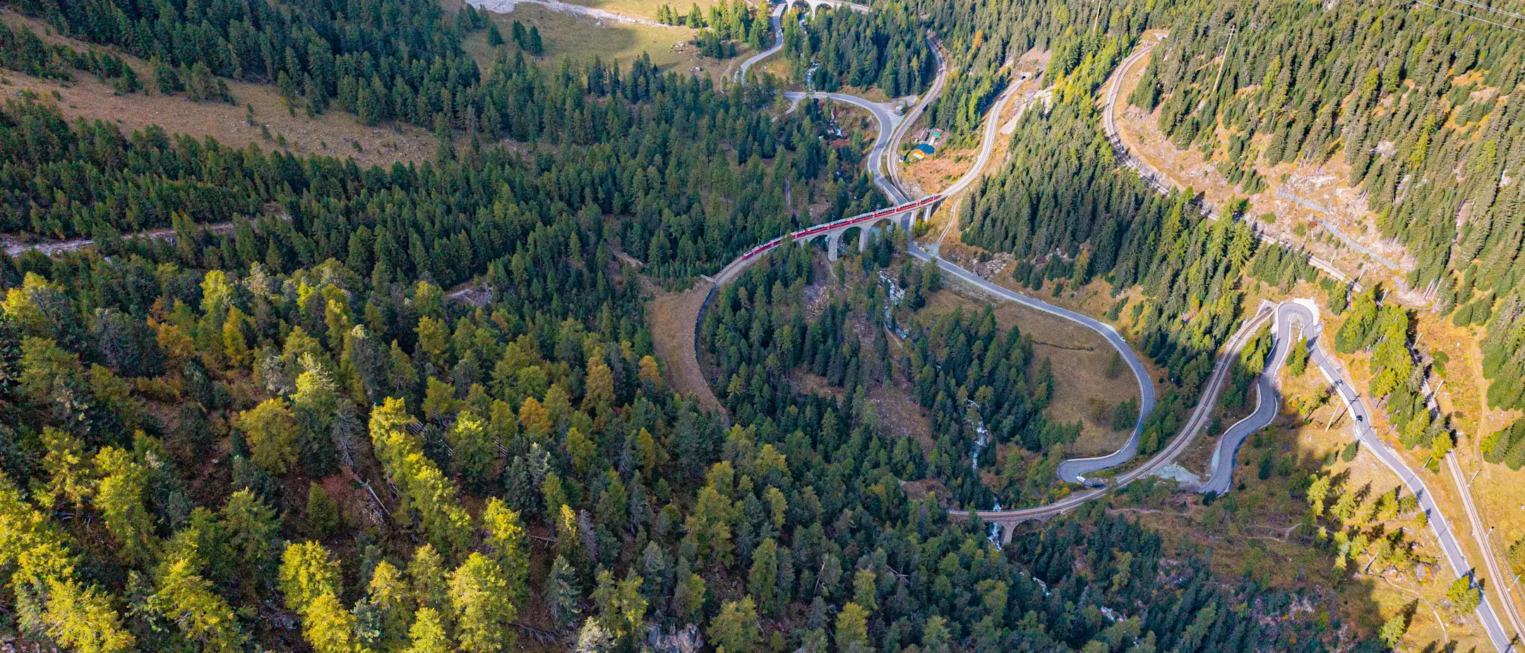 Luftaufnahme einer spektakulären Bahnstrecke, die sich in eleganten Kurven und Tunneln durch ein bewaldetes Tal windet.