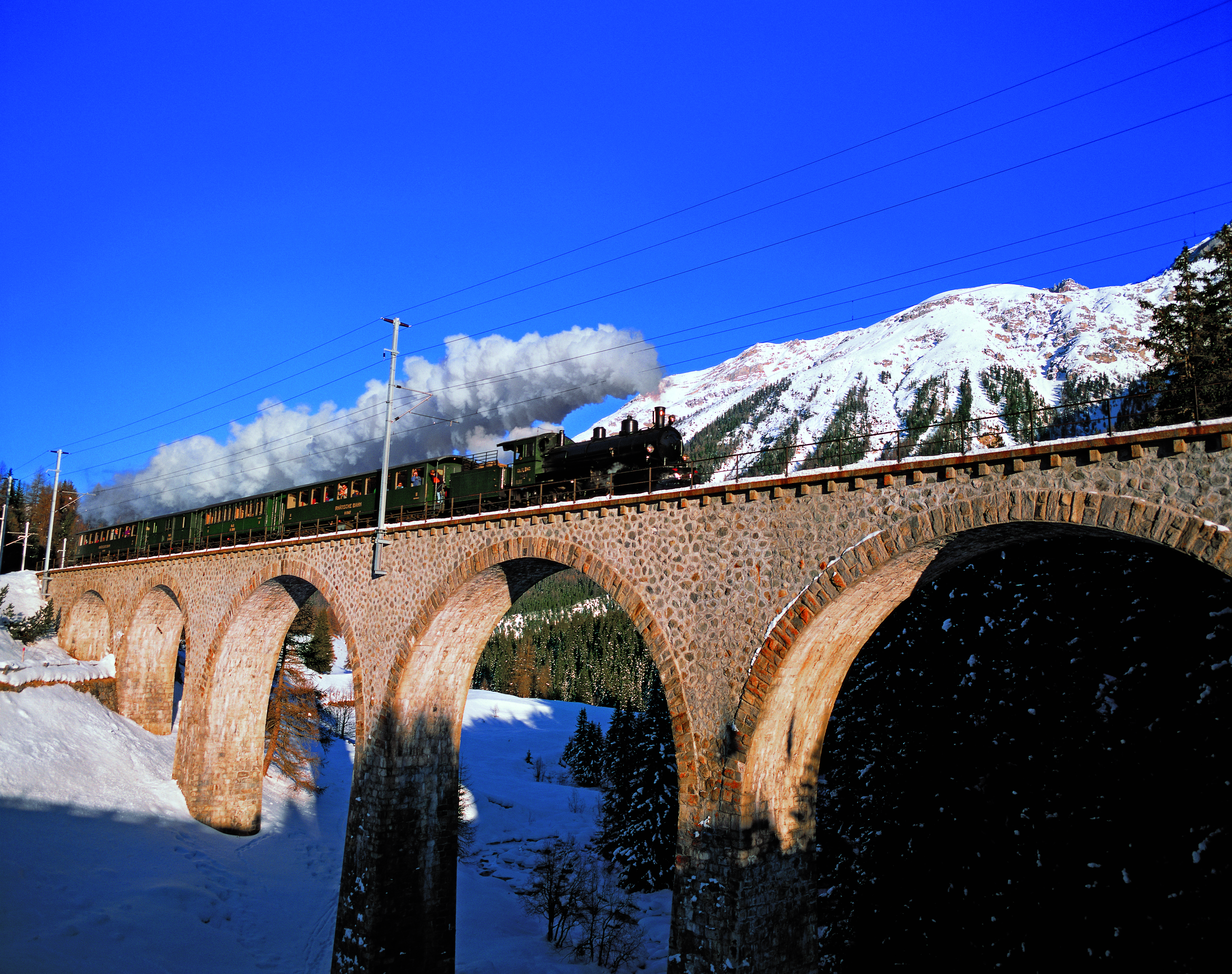 Dampfzug der Rhätischen Bahn überquert im Winter das Viadukt bei Cinuos-chel, schneebedeckte Berge im Hintergrund.