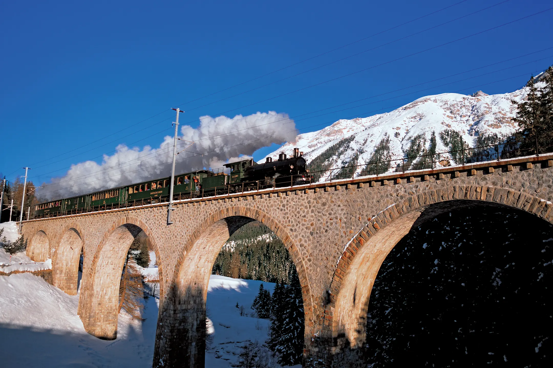 Dampfzug der Rhätischen Bahn überquert im Winter das Viadukt bei Cinuos-chel, schneebedeckte Berge im Hintergrund.