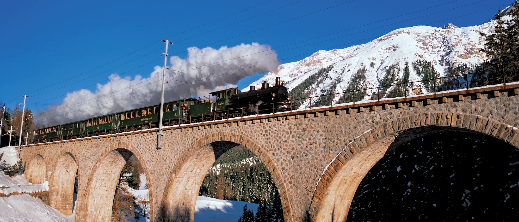 Dampfzug der Rhätischen Bahn überquert im Winter das Viadukt bei Cinuos-chel, schneebedeckte Berge im Hintergrund.